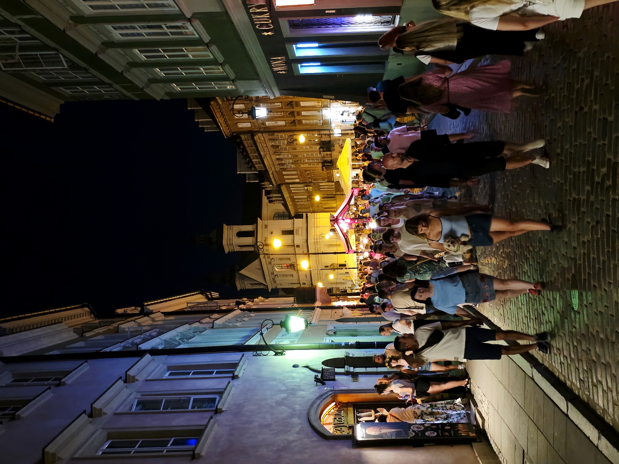 The image depicts a crowded cobblestone street at night in Warsaw, Poland, likely within the Old Town or a similar historic district. The street is lined with old, European-style buildings with ornate facades, illuminated by streetlights and building lights. A significant number of people are walking along the street, suggesting a lively atmosphere, possibly a market or evening promenade.
On the left side of the street, a building with a pale pink facade features arched doorways and windows. One doorway has a sign that appears to read "CZYTAJ" in large letters, and below it, a display featuring images of ice cream. Another sign on the wall reads "20.94". A green streetlamp is mounted on the building.
The right side of the street is dominated by buildings with darker facades, one of which has illuminated signage reading "* WINA" and "* CUKRY". The street is paved with cobblestones, and shadows are cast by the people and buildings.
The people in the image are diverse in age and attire, with many dressed in casual summer clothing. Some are walking in groups, while others are alone or in pairs. One woman in the foreground on the right is holding a small dog. The overall impression is of a vibrant urban scene at night, with a mix of architectural styles and a lively pedestrian presence.