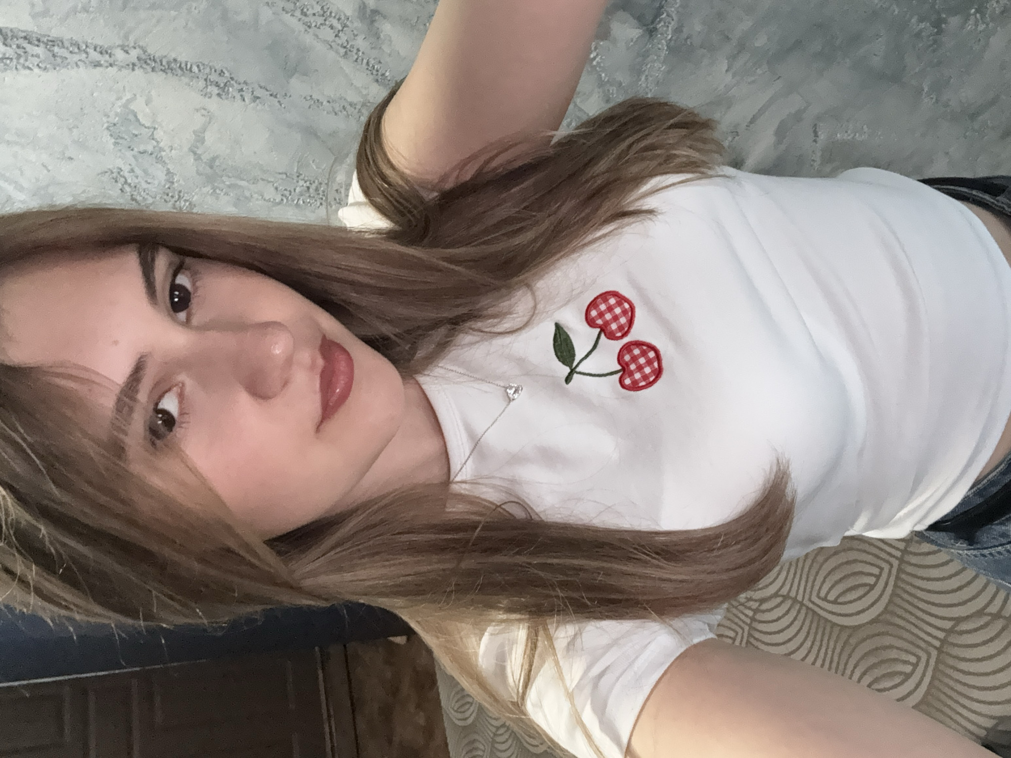 A young woman is depicted in a close-up, horizontally oriented selfie, likely taken while lying down indoors in Panagyurishte, Bulgaria. She has long, straight brown hair that cascades over her shoulders and chest. Her face is turned towards the camera, showing a neutral to slightly pleasant expression with her dark eyes looking directly at the viewer and lips lightly tinted.
She is wearing a white t-shirt adorned with a distinctive embroidered design of two red and white checkered cherries, complete with a green stem, on the left side of her chest. A delicate silver necklace with a small, shimmering heart-shaped pendant hangs around her neck. The lower part of her body is partially visible, suggesting she is wearing dark blue jeans or similar attire.
The background includes a light-colored, textured wall behind her head, possibly a patterned wallpaper in hues of light blue and grey. Below her body, another patterned surface with swirling, light-colored lines on a beige background is visible, likely a bedspread or upholstery. The indoor lighting appears natural, suggesting it is daytime. There is no visible text in the image.