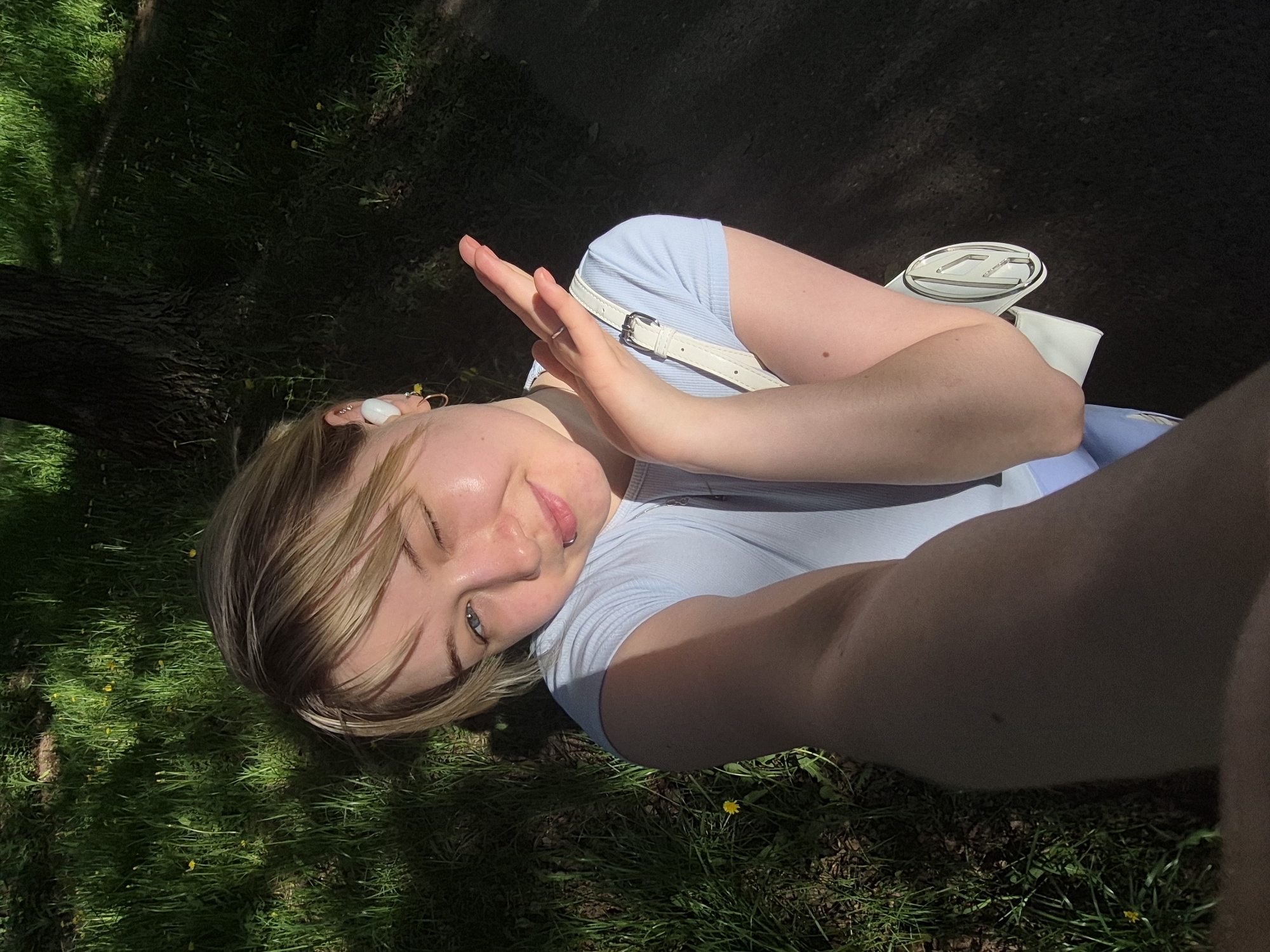A young woman takes a selfie outdoors on a sunny day. She is fair-skinned with blonde and brown hair, and one eye is closed in a wink. She is wearing a light blue t-shirt and has a white purse slung over her shoulder. Her hands are raised as if blocking the sun. The location is a grassy area with trees, likely a park. Given the prompt, the location could be within the city of Chaikovsky, Russia. The ground is covered in grass with some yellow flowers. Part of a tree trunk and some dark shadows are visible in the background, along with a path.