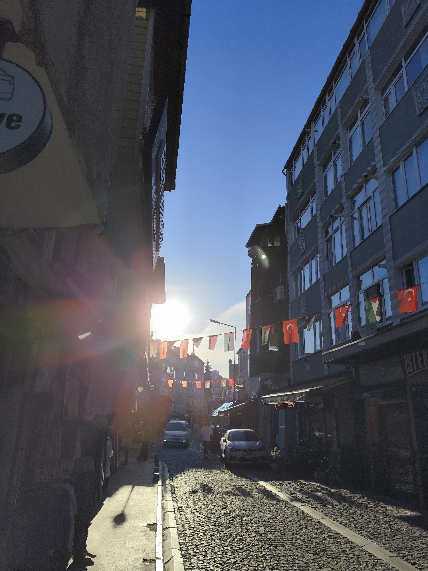 The image shows a street in Sarıyer, Turkey, illuminated by bright sunlight, creating stark contrasts of light and shadow. The street is paved with cobblestones and lined with buildings of varying heights, one of which has flags of multiple countries strung between them. Several people are walking along the street, and two cars are parked along the curb. A business sign is visible on the left.