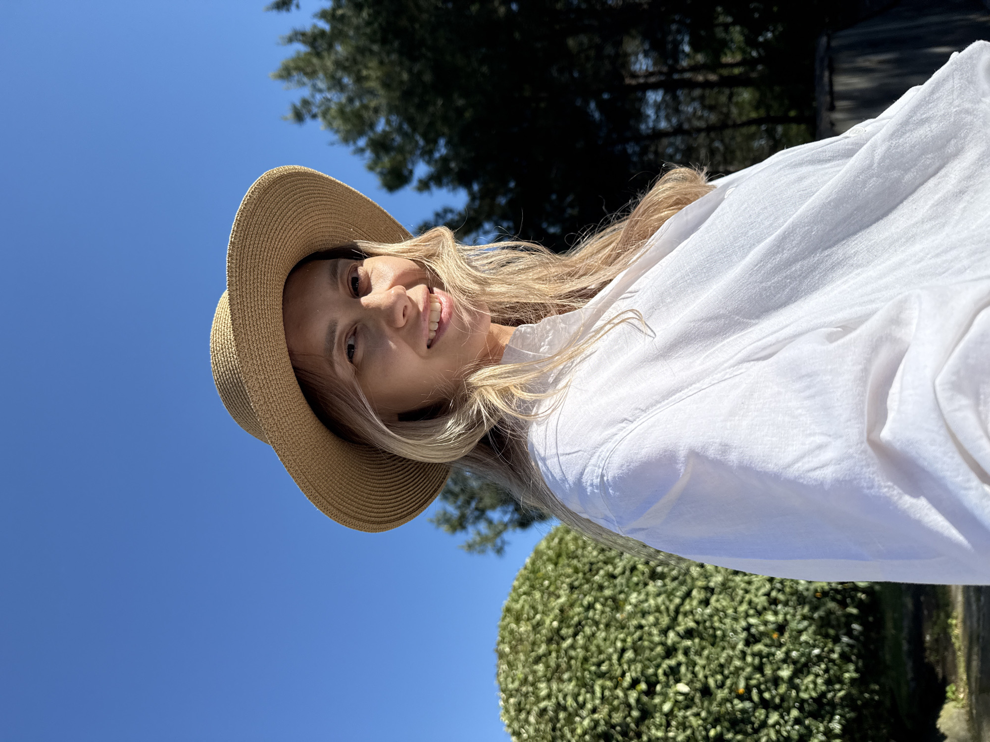 A bright, outdoor photograph, taken in Nebug, Russia, features a smiling woman with long blonde hair, wearing a straw hat and a white long-sleeved shirt. The image is rotated, but depicting the woman looking upwards towards the viewer from a slightly low angle. Her face is cheerful, illuminated by direct sunlight, highlighting her smile.
The background is dominated by a clear, vibrant blue sky, suggesting excellent weather. Below the horizon line, dark green trees are visible in the distance, and to the right, a large, well-maintained, spherical green bush adds a touch of natural beauty to the scene. The lighting indicates a sunny daytime, with no clouds visible. The woman's casual attire, including the straw hat, suggests a relaxed, possibly vacation-like, setting. No text is visible in the image.
