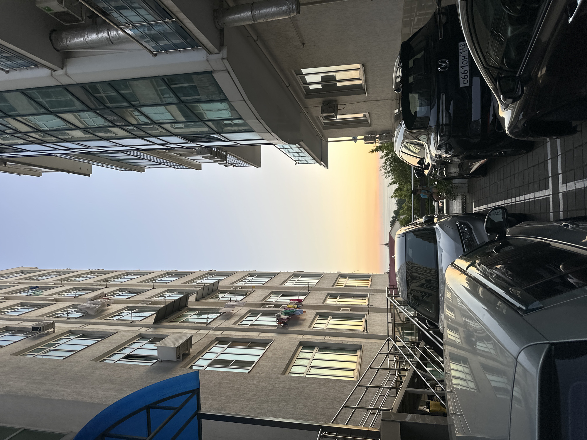 A low-angle shot captures an urban scene with two multi-story buildings flanking a narrow alleyway. The building on the left, constructed from concrete, features numerous rectangular windows, some with air conditioning units installed below them. Laundry is visible drying on a clothesline strung between windows on one of its facades. A blue awning shades a section of the building near its base, where metal stairs lead upwards. The building on the right has a modern facade dominated by large, reflective glass panels, with balconies projecting outwards. Several cars are parked in designated spots within the alley, including a silver SUV, a black SUV, and a dark-colored sedan. The scene is bathed in the warm light of a sunset, with the sky displaying hues of orange and yellow, transitioning to a clear blue overhead. In the distance, a body of water, likely the Black Sea, is visible beyond a line of trees and some distant buildings. A few individuals can be seen on a balcony overlooking the sea. The overall impression is one of an apartment complex in a coastal city, possibly Sochi, Russia, during the evening golden hour.
