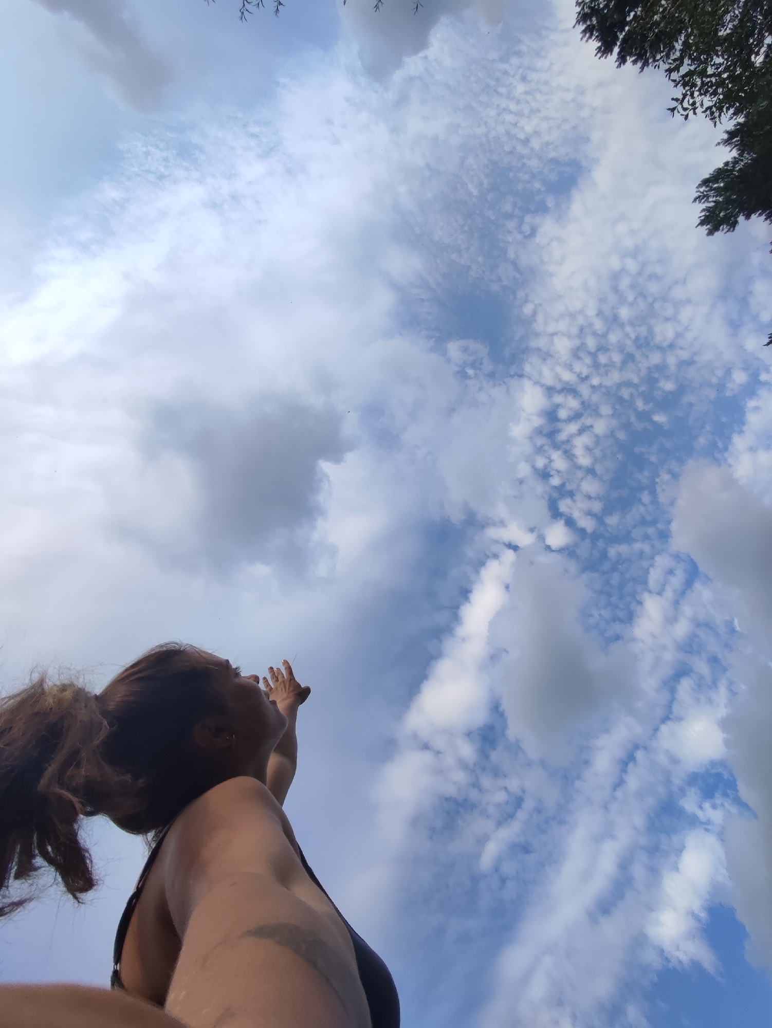 A low-angle, full-shot photograph captures a woman from the chest up, looking up at a cloudy blue sky. Her brown hair is pulled back into a ponytail that cascades over her left shoulder. Her face is turned to the right, with her mouth slightly open as if speaking or reaching for something. Her right arm is extended upwards, with her hand open and reaching towards the sky. She is wearing a dark, possibly black, tank top. Visible on her left arm is a dark, curved tattoo. The sky is filled with a mix of white and gray clouds, with patches of blue sky visible. The top corners of the image show the dark green foliage of trees. The image does not provide any specific details about the location within the city of Фоки, Russia.