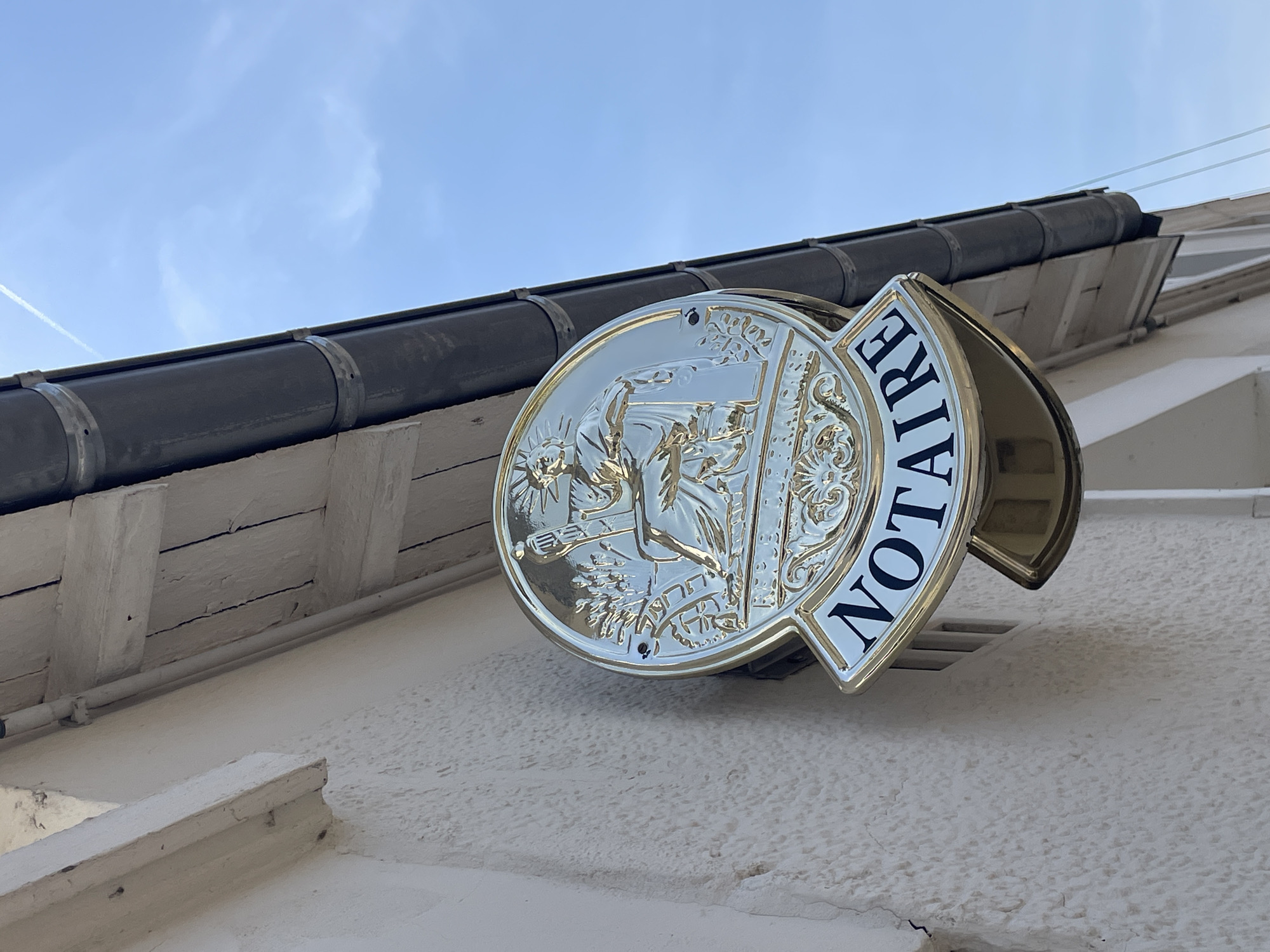 The image shows a close-up, eye-level shot of a gold-colored circular sign with the word "NOTAIRE" in black lettering on a white background, attached to the exterior wall of a building. The sign features an embossed image of a robed figure holding a staff and scales, a common symbol for justice or legal professions. The building's wall is textured and painted white, and a dark gray gutter runs along the roofline above the sign. The sky above is a clear blue with faint white contrails from an airplane. The scene appears to be an exterior view of a professional office in Meaux, France, likely a notary's office given the signage.