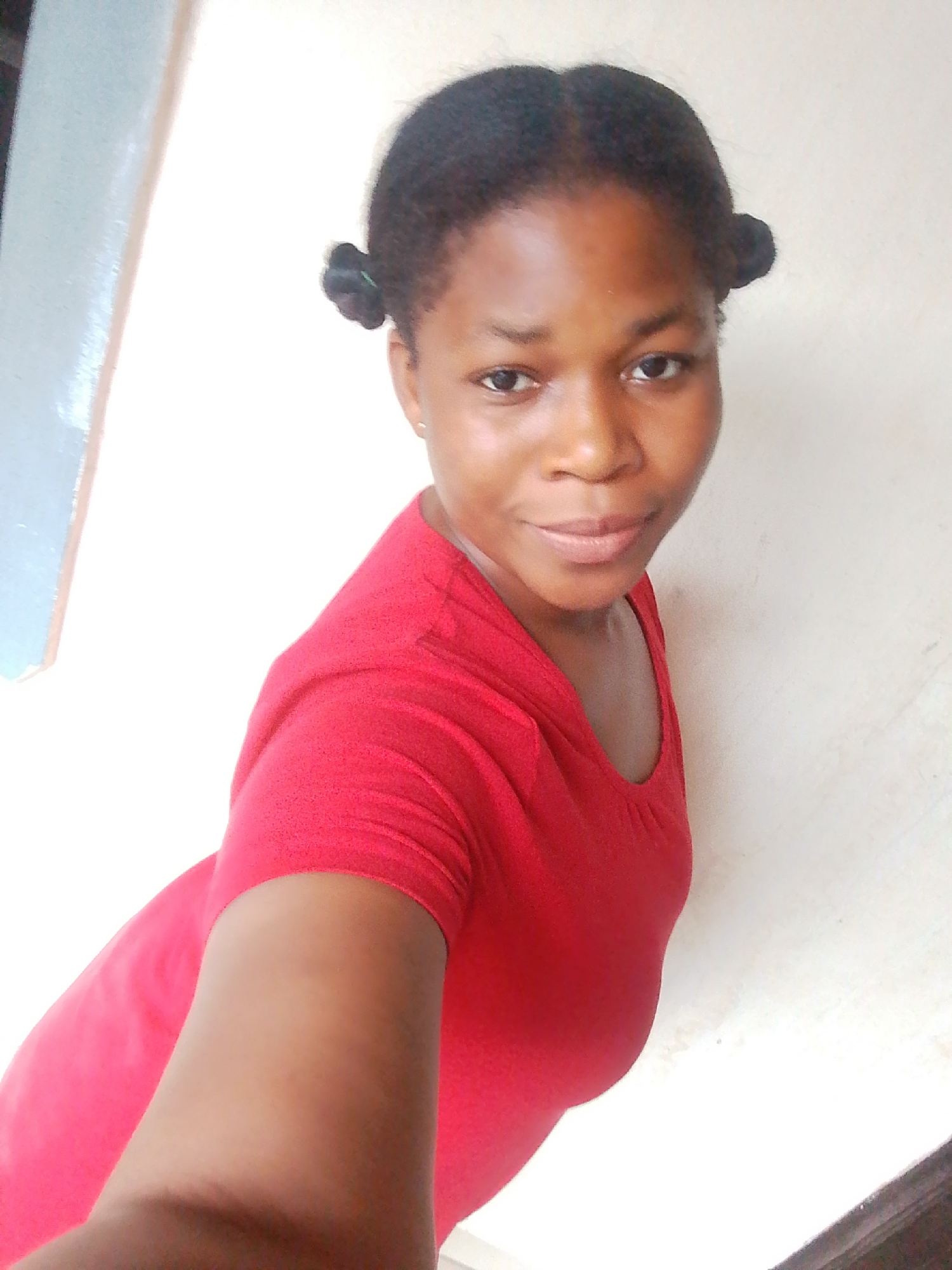 A young Black woman is pictured in a selfie, looking towards the camera with a slight smile. She has her hair pulled into two small buns on the sides of her head. She is wearing a red t-shirt. The background is a plain, light-colored wall, with a hint of a blue-painted surface to the left of the frame, suggesting an indoor setting or a covered outdoor area. The lighting appears to be natural, possibly from daylight. The woman's expression is relaxed and friendly. There is no visible text in the image. The setting could be a home or a similar informal environment in Yenegoa, Nigeria. The image captures a casual moment.