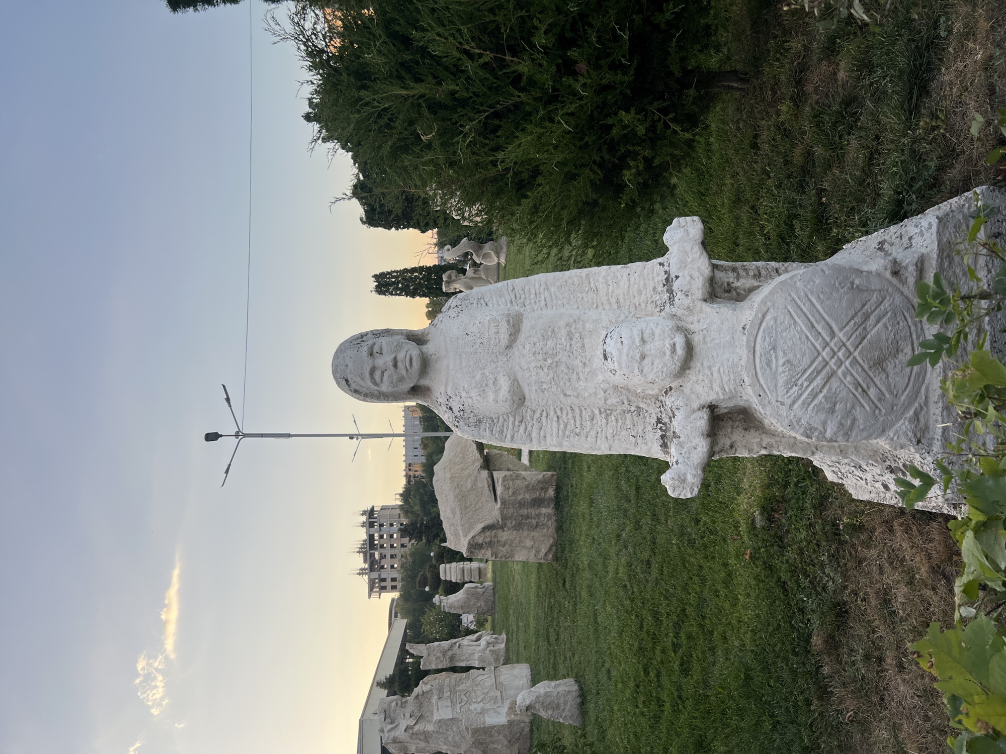 In Osh City, Kyrgyzstan, a statue of a woman and a child stands in a grassy outdoor area. The statue, made of stone, depicts the woman with her arms around the child, who is holding a circular shield. The scene appears to be in a park or sculpture garden, with other stone sculptures and lush green trees surrounding the main subject. In the background, a multi-story building under construction is visible, along with a street lamp. The sky above is clear with a hint of clouds, suggesting it is either early morning or late afternoon.