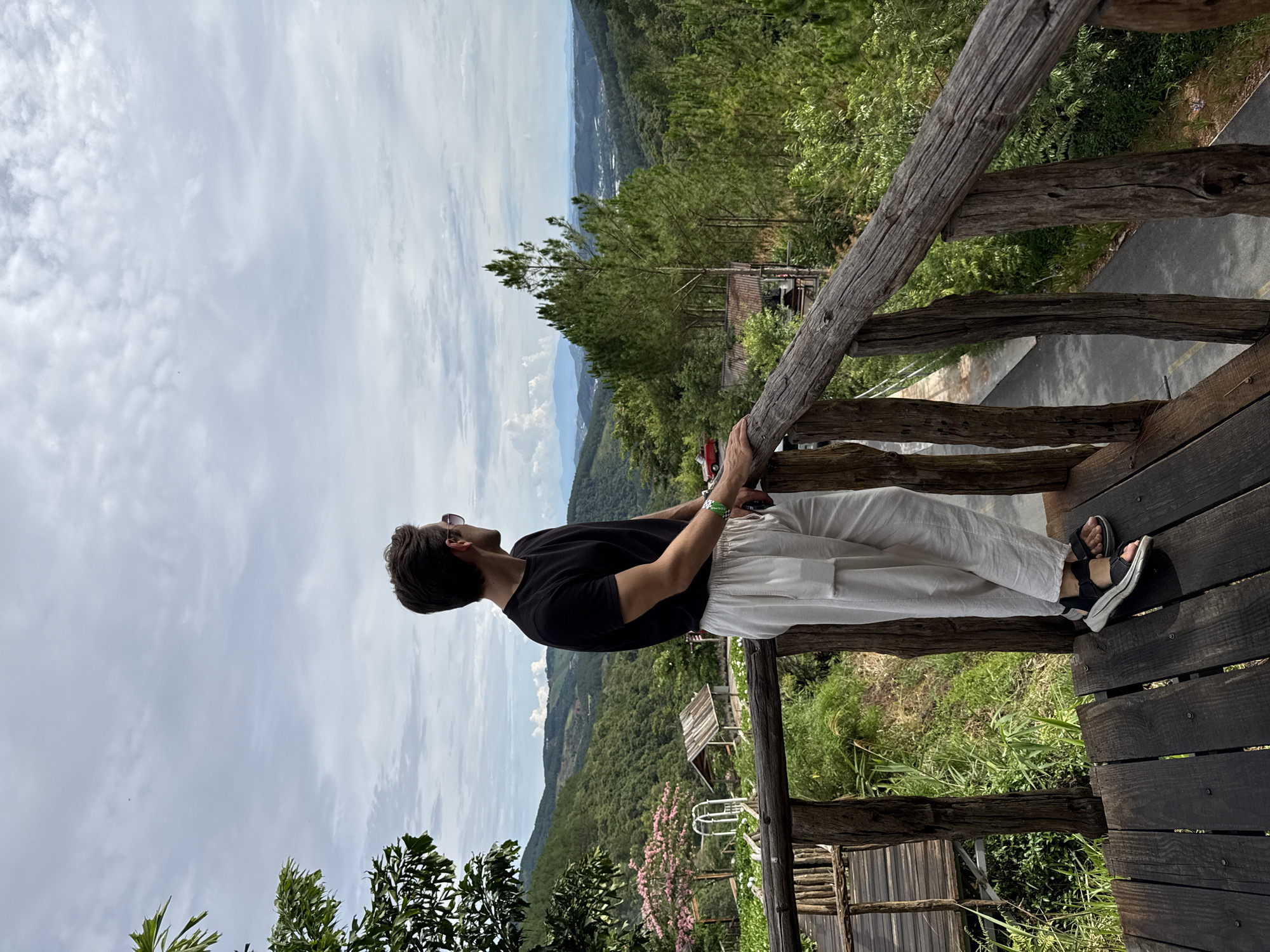 A person stands on a wooden platform overlooking a mountainous landscape under a cloudy sky. The person is wearing a black t-shirt, white pants, and sandals. They are leaning against a wooden railing, looking out at the view. The background shows rolling hills covered in trees, with patches of farmland and distant buildings visible. The wooden platform appears to be part of a structure built on the side of the mountain in Thôn 5, Vietnam.