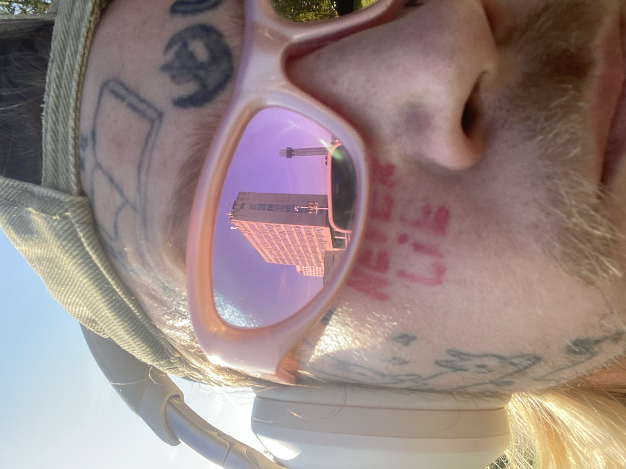 This is a close-up, low-angle, portrait-style photograph of a person's face wearing pink-tinted sunglasses. The person appears to be outside, as indicated by the bright sunlight and a clear blue sky visible in the upper left quadrant of the image. The sunlight casts a bright glare on the subject's forehead and glints off the sunglasses.
The subject has several tattoos on their face and forehead, including what appears to be a rectangular shape with lines inside, a silhouette of a cat or similar animal, and the text "NEVER LIE" in red script on the cheek. The person is also wearing a light-colored, possibly canvas or denim, hat and headphones around their neck. The reflection in the sunglasses shows a tall, brick building with many windows and a structure resembling a water tower or chimney on top. The reflection is tinted purple, likely due to the lens color.
The framing of the shot is unusual, with the camera positioned below the subject and looking upwards. This perspective, combined with the close-up nature of the photo, emphasizes the subject's face and the details of their tattoos and accessories. The overall mood is somewhat candid and possibly defiant, given the "NEVER LIE" tattoo. The time of day appears to be daytime, with the sun high in the sky. There are no overt indications of the specific location beyond what can be inferred from the architecture reflected in the glasses.