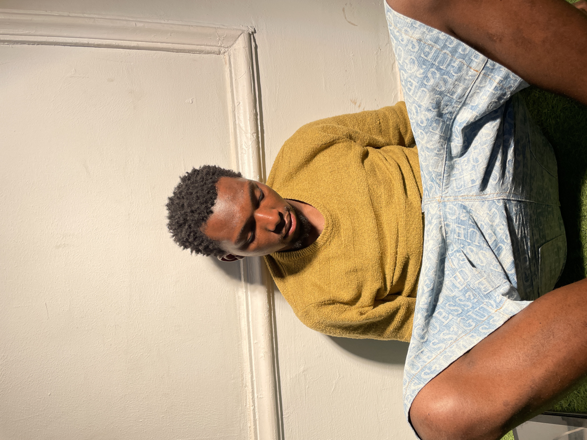 A medium shot depicts a Black man lying in a relaxed pose. The image is oriented horizontally, with the man's head positioned towards the left and his lower body towards the right, appearing to rest against a light-colored wall with architectural molding visible along the top edge of the wall. A textured green surface, possibly artificial grass or a rug, is visible beneath his legs on the right side of the frame.
The man has dark skin and short, curly black hair. His eyes are closed or partially closed, suggesting a state of relaxation or sleep. He is dressed in a mustard yellow long-sleeved knit sweater and light blue shorts. The shorts feature a repeating pattern of the word "GUESS" printed in white.
Strong, natural light from the top-left (relative to the current image orientation) illuminates the scene, creating distinct shadows on the man's face, clothing, and the wall. This indicates a bright, sunny daytime environment. The wall itself has a slightly textured surface and some minor blemishes. The overall mood conveyed is peaceful. The image was taken in Barakin Akawo, Nigeria.