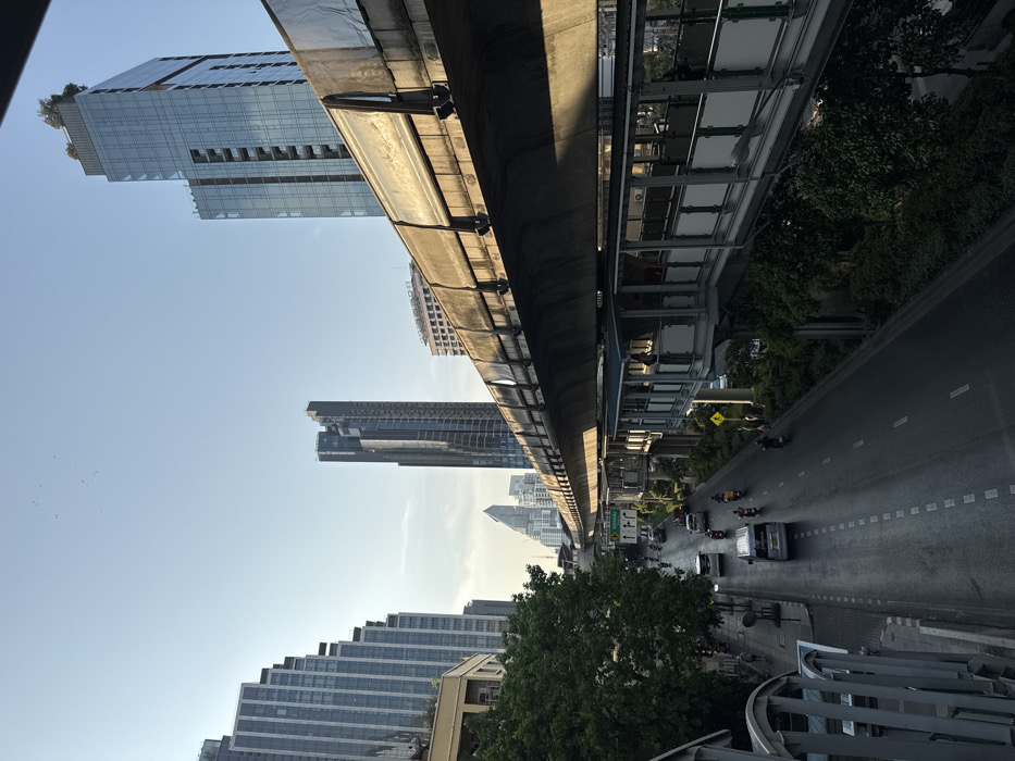 The image displays an urban scene in Bangkok, Thailand, situated along Ratchadamri Road near the Ratchadamri BTS Station, viewed from an elevated perspective looking south-east. The sky is clear and light blue, with a small flock of birds visible in the upper-left.
A prominent concrete elevated railway track, identified as part of the BTS Skytrain infrastructure, traverses the middle and right portions of the frame, casting a large shadow over the roadway and a lower pedestrian structure. The track exhibits weathered surfaces and some discoloration. Beneath a section of this railway, a secondary elevated pedestrian walkway, constructed from metal and glass with a light blue roof, runs parallel. Several individuals are present on this walkway; one person stands at the railing on the right, while others are seen walking or standing further along.
Below the elevated structures, a wet, multi-lane asphalt road extends into the distance, marked with white dashed lines. Traffic includes a light-colored sedan, a silver van, and multiple motorcycles, moving in both directions. Pedestrians are visible on the sidewalks, particularly on the left side, which is lined with green trees. An overhead green traffic sign structure is discernible on the left.
The background features a dense collection of modern skyscrapers. On the far right, a tall, glass-clad building with a stepped facade and rooftop greenery is prominent, identifiable as Magnolias Ratchadamri Boulevard. To its left, another very tall, dark-colored skyscraper with a complex, segmented architectural design stands further in the background. On the far left, a series of high-rise buildings with light-colored, rectilinear facades are visible.