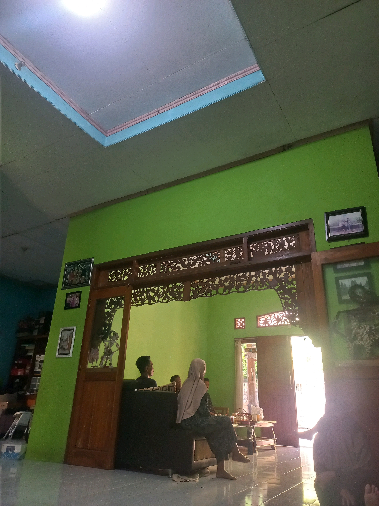 The image depicts the interior of a residence in Sukoharjo, Indonesia. The room has bright green walls and a light-colored ceiling with decorative trim. In the foreground, a wooden doorway with intricate carvings serves as an entrance to another part of the house. Several framed photographs are displayed on the green wall to the left of the doorway.
To the right of the doorway, another wooden-framed opening reveals a glimpse of an outdoor area with sunlight streaming in. Inside the room, a few individuals are present. In the center, a woman wearing a light-colored hijab and patterned clothing is seated on what appears to be a couch or bench. Next to her, a person, possibly a man, is also seated. Further to the right, partially obscured by shadow and the edge of the frame, is another individual, possibly a woman, with bare feet visible. A low table with some items on it is situated in front of the seated individuals. The floor is tiled and reflects the light. The overall impression is a casual indoor setting, likely a living area.