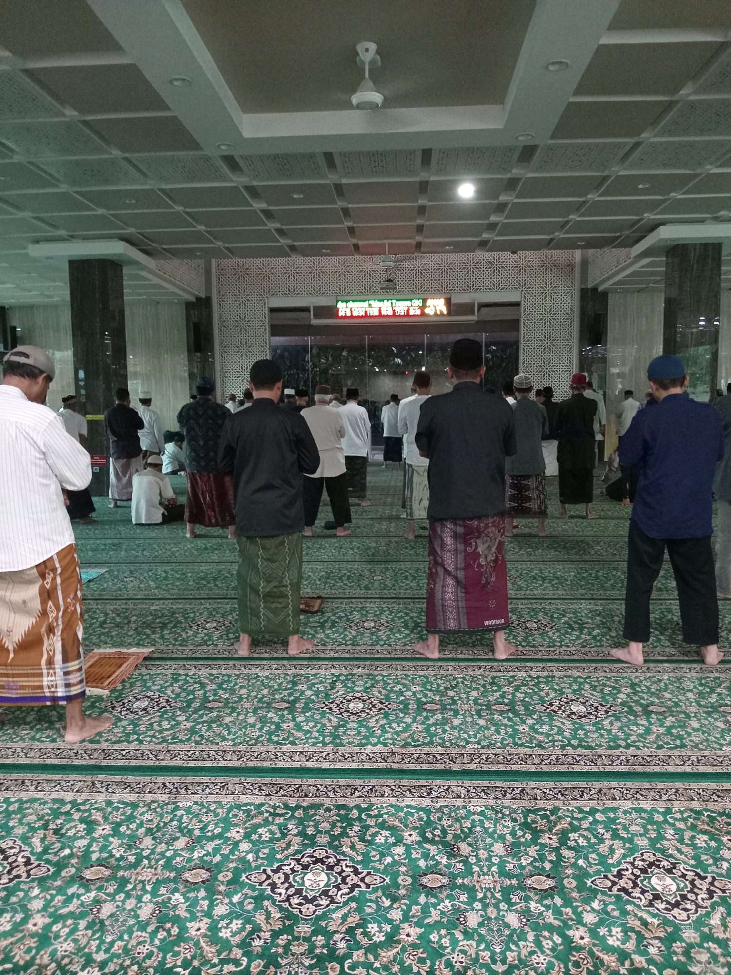 The image depicts the interior of a mosque, likely located in Randuagung, Indonesia, judging by the context. Several men, mostly wearing sarongs and traditional tops, are standing in rows facing what appears to be the front of the mosque, suggesting they are engaged in prayer or waiting for prayer to commence. The mosque features a green carpet with an intricate pattern, columns, a patterned ceiling with lighting fixtures and a ceiling fan, and a large screen displaying red text in a foreign script. The lighting is artificial, casting a soft glow on the scene. The overall atmosphere is one of communal religious gathering.