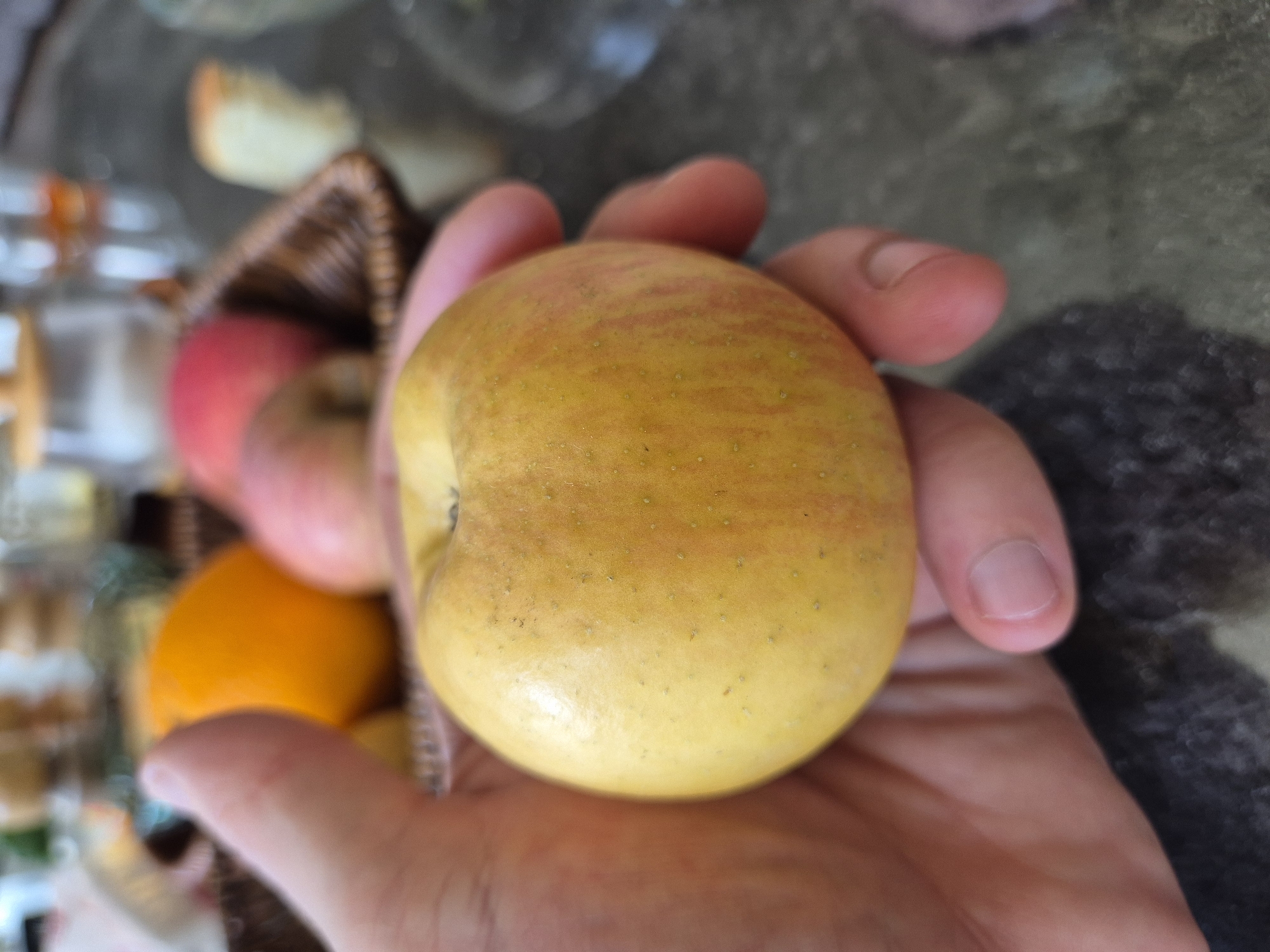 A close-up, eye-level shot focuses on a person's hand holding a yellow and red streaked apple. The apple is positioned in the center of the frame, filling much of the space. In the background, blurred fruits, including an orange and a red apple, are visible within a wicker basket. The surface below the basket appears to be a dark, reflective tabletop. The overall scene suggests a casual, possibly domestic setting. There is no visible indication of the city of Taldykorgan, Kazakhstan.