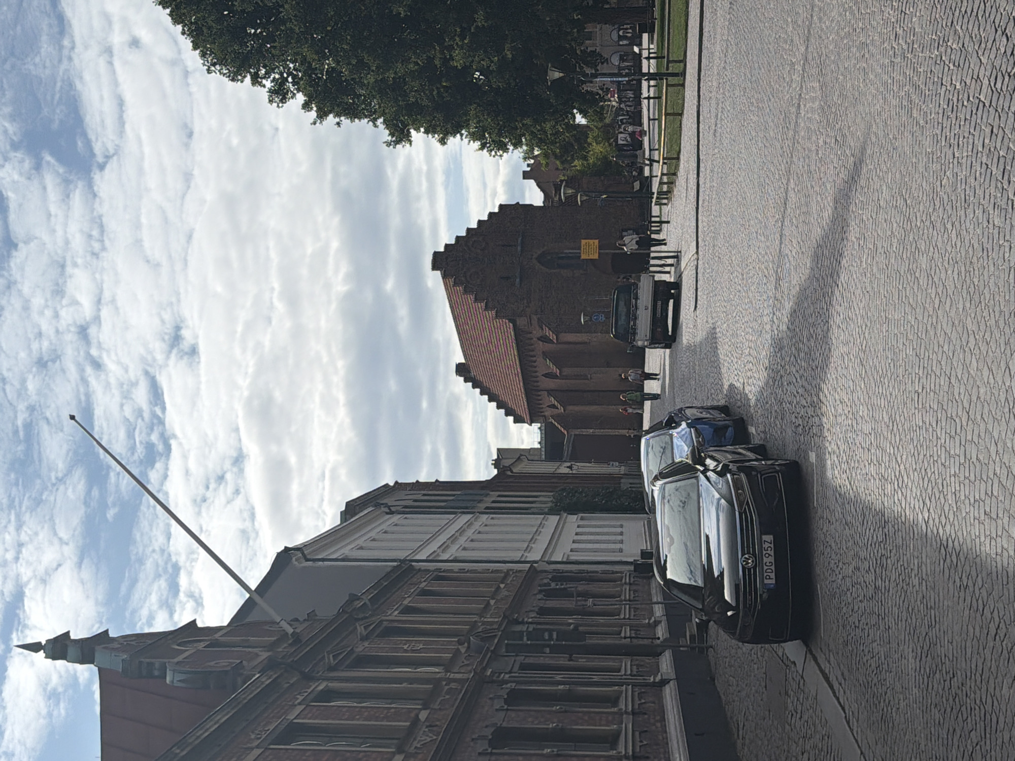 The image captures a street scene in Lund, Sweden, featuring historic architecture and urban elements under a partially cloudy sky. On the left, a row of old brick buildings with detailed facades lines the cobblestone street. A black Volkswagen sedan, license plate PDG 95Z, is parked closest to the camera, followed by a blue car. In the background, a distinct brick building with a red tiled, stepped gable roof and arched windows stands prominently. Several pedestrians are visible walking on the sidewalk near this building and further down the street. A white pickup truck is parked on the right side of the street. A large, dark green tree dominates the right side of the frame, casting a shadow on the cobblestone pavement. A tall flagpole extends diagonally across the upper portion of the sky, originating from a building on the left.