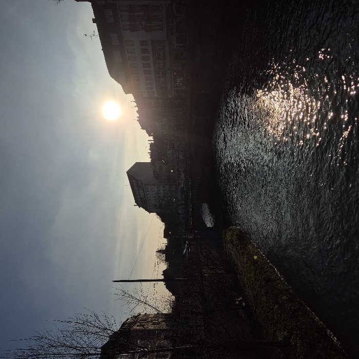 The image displays an outdoor scene, rotated 90 degrees counter-clockwise, featuring a body of water, buildings, and a low sun. If viewed correctly oriented, the scene depicts a river or canal flanked by urban structures, bathed in the warm light of what appears to be either sunrise or sunset.
In the upper central portion of the correctly oriented image, a bright, circular sun shines through a slightly hazy sky, casting a strong, warm glow. The sky around the sun is light, transitioning to a cooler blue-grey towards the upper left. The sun's intense light creates silhouettes of the buildings and objects directly in its path.
To the right of the sun, a body of dark water, likely a river or canal, flows. The surface of the water is heavily textured with ripples and reflects the sunlight in numerous sparkling points, creating a shimmering effect. The reflections are particularly intense closer to the sun's position.
Along the left bank of the water (upper part of the rotated image), a row of multi-story buildings is silhouetted against the bright sky. Their facades are mostly dark, but some architectural details, like windows and rooflines, are subtly visible, hinting at traditional European building styles. Another building or complex of buildings is visible further back on this bank, also appearing dark against the light.
On the right bank of the water (lower part of the rotated image), the shoreline features a dark, mossy or vegetated stone wall that extends into the foreground. Beyond this wall, the bank is lined with dark, indistinct vegetation and bare tree branches are visible in the lower right foreground. A tall, slender pole, possibly a utility pole or lamppost, stands on this bank.
The overall impression is one of dramatic contrast between the brilliant sun and its reflections, and the dark, silhouetted forms of the city and landscape. No specific text is visible in the image. The scene evokes a quiet, possibly late afternoon or early morning atmosphere.