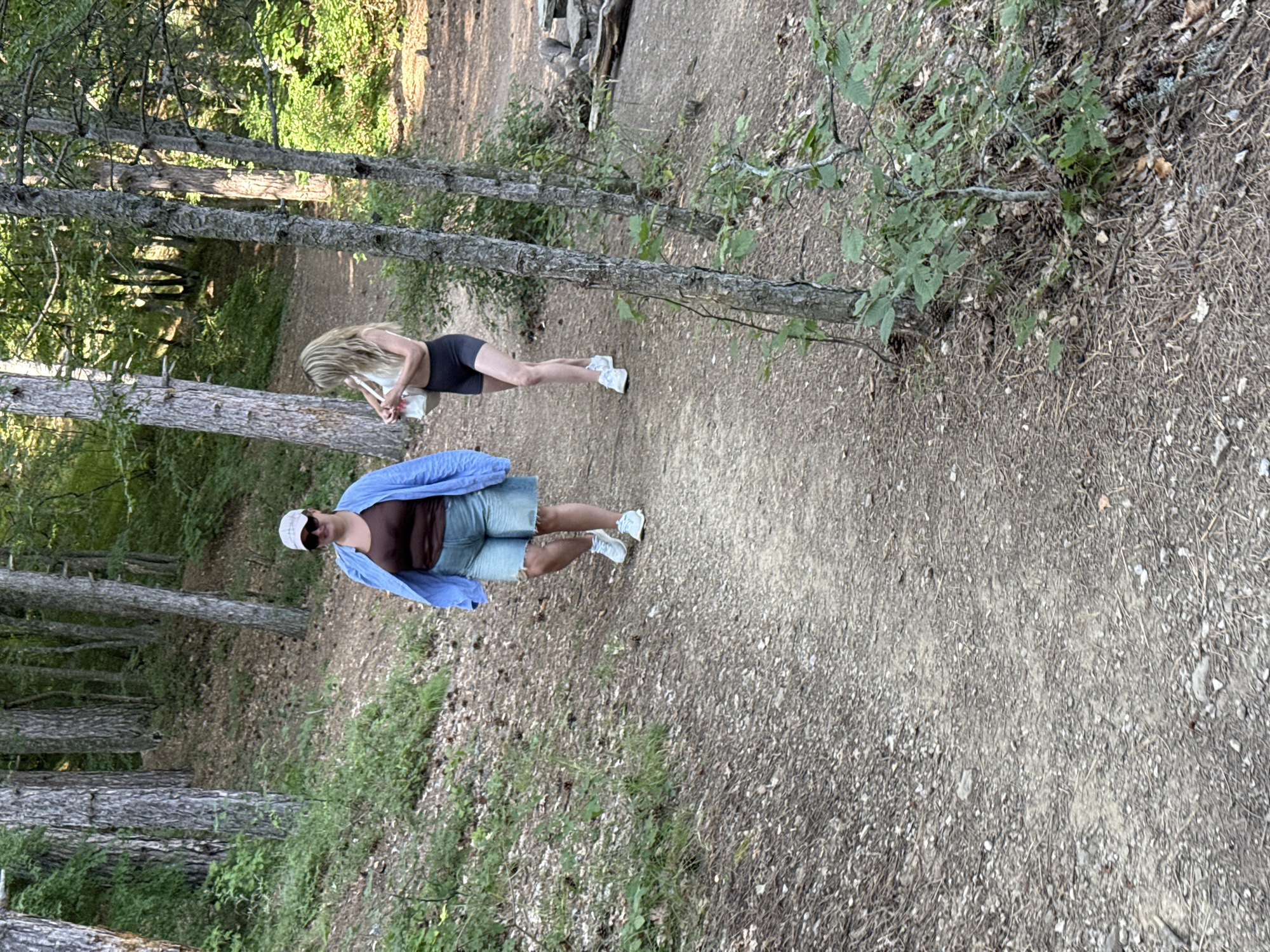 Two individuals are walking on a dirt path in a forest. The person in the foreground is wearing a brown tank top, a blue unbuttoned shirt, and denim shorts, along with a white baseball cap. The second person, in the background, is wearing a white crop top and black shorts, carrying a white bag. The path is surrounded by trees and vegetation. There are some rocks and logs visible to the right of the path. The scene suggests an outdoor recreational activity. The location is identified as Zaprudnoe, Ukraine.