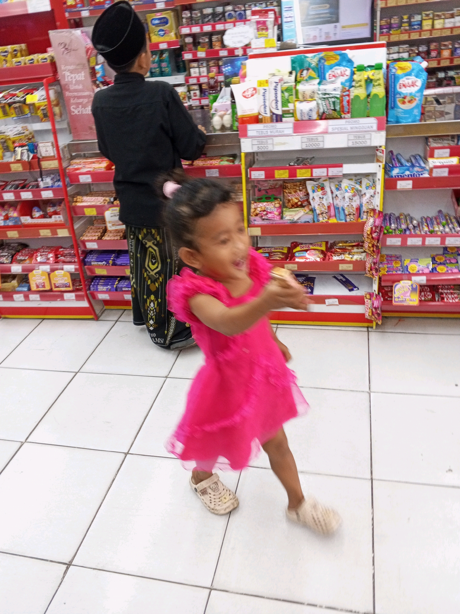 In a brightly lit convenience store in Ciruas, Indonesia, a young girl wearing a vibrant pink dress and beige clogs is captured mid-motion, her right arm extended as if reaching for something or in the process of throwing an object. Her face is blurred due to motion, but her open mouth suggests excitement or speech. Behind her, a person in traditional attire, a black peci cap and a black shirt over a batik sarong, stands facing away from the camera, possibly a parent or guardian. The background is filled with shelves stocked with various packaged goods, including snacks, drinks, and household items, typical of a retail environment. The floor is tiled with large white squares, and the overall impression is one of a busy, everyday scene.