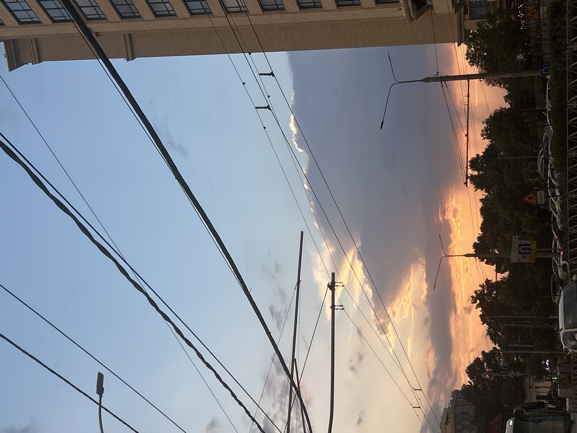 The image captures a sunset scene in Krasnodar, Russia. The sky is a gradient of blue and orange, with dark clouds obscuring parts of the sunset. Overhead, a complex network of power lines crisscrosses the frame, with a street lamp visible on the right. A tall, modern building with multiple windows stands to the right of the frame, and a lower building can be seen in the background on the left. Below the power lines, the tops of trees form a dark silhouette against the colorful sky. Further down, a street scene unfolds with more trees, street lamps, road signs, and parked cars. The overall impression is of an urban environment bathed in the warm, dramatic light of a sunset.
