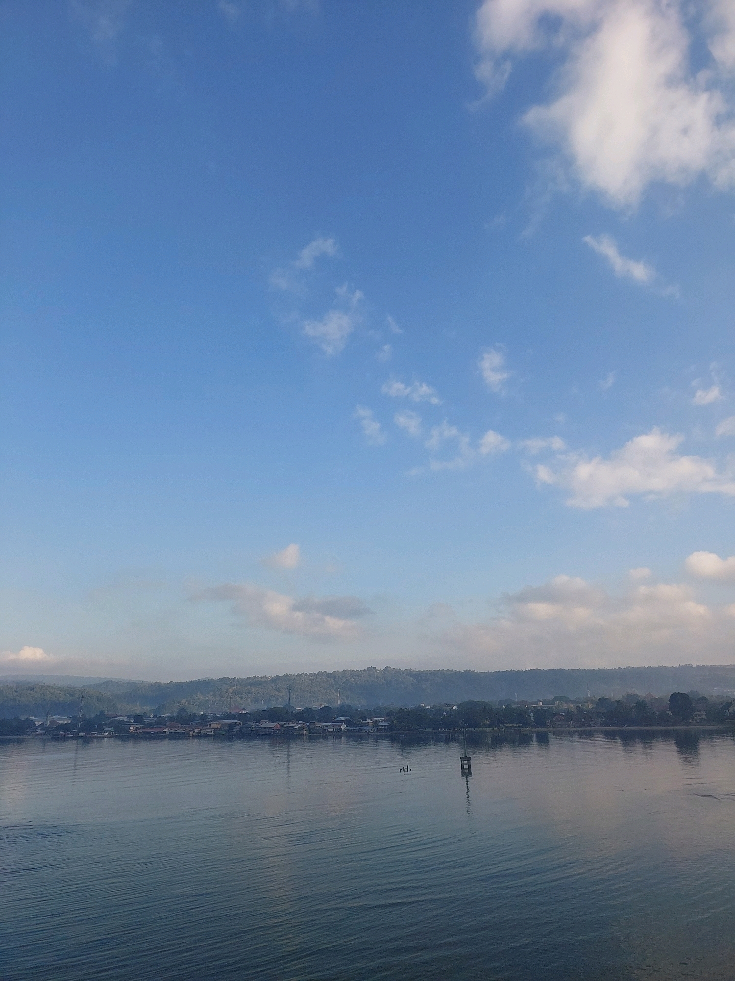 The image depicts a serene waterfront scene in Ambon, Indonesia. A vast expanse of calm, rippling water occupies the foreground, reflecting the blue sky and scattered white clouds above. In the middle ground, a long, low-lying coastline is visible, characterized by lush green foliage that gives way to a dense settlement of buildings. The architecture suggests a tropical setting, with many structures appearing to be built on stilts or close to the water's edge. A solitary, weathered wooden structure, possibly a navigation marker or part of a pier, stands in the water, breaking the monotony of the calm surface. In the distance, rolling hills covered in trees form a natural backdrop to the urban landscape. The overall impression is one of tranquility and a harmonious blend of nature and human habitation, typical of coastal communities in this region.
