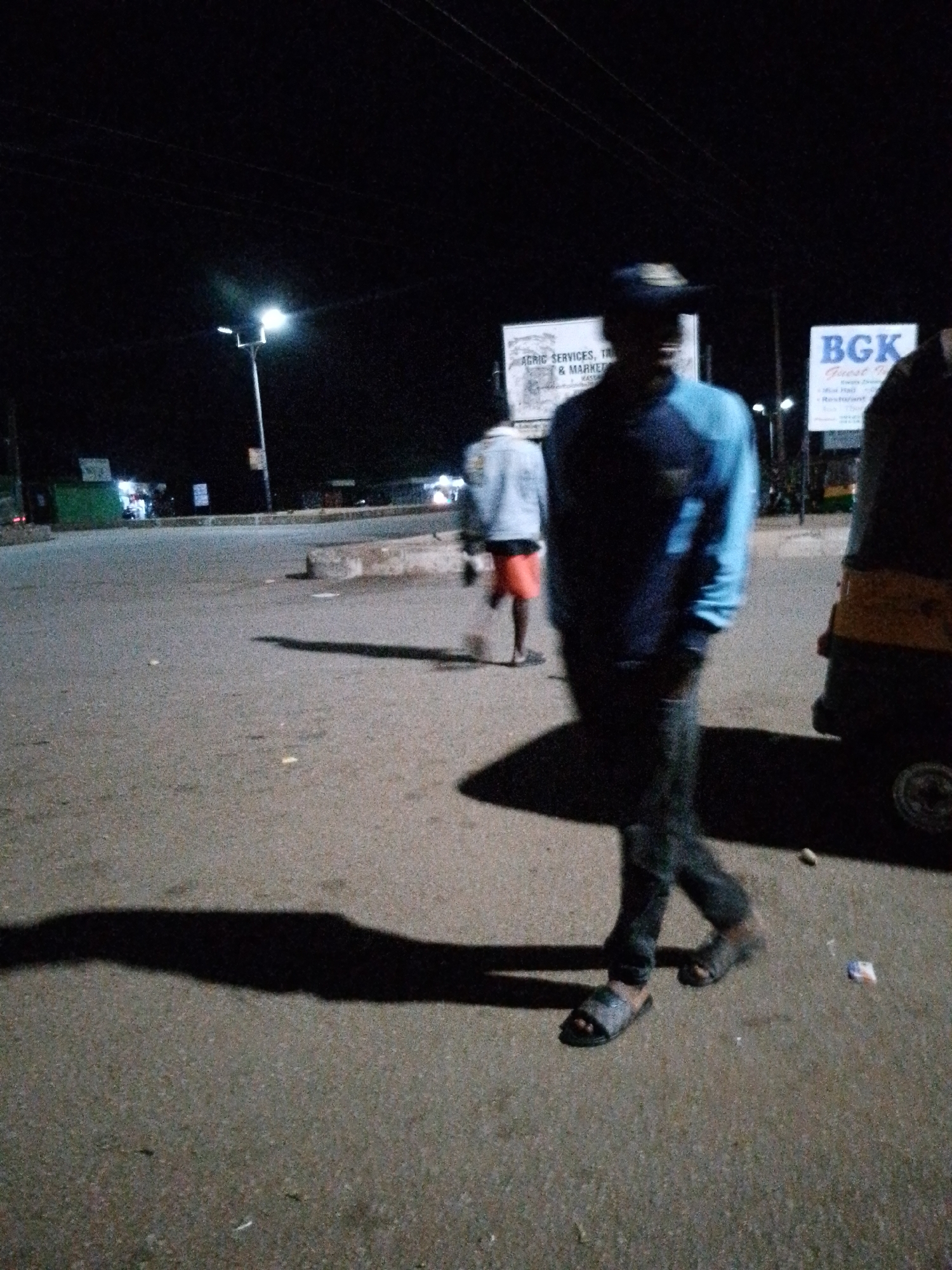 In Bukuru, Nigeria, at nighttime, two individuals are present in a sparsely populated urban area. A man in the foreground, wearing a blue long-sleeved shirt, dark pants, and sandals, walks to the right, casting a prominent shadow. Behind him, another person, wearing a light-colored long-sleeved shirt and orange shorts, walks away from the camera. The environment appears to be a paved road or open area, possibly a transportation hub, with streetlights providing illumination. Signs with text are visible in the background, including one that reads "BGK". A portion of a three-wheeled vehicle is visible on the right side of the frame. The overall scene is characterized by low light conditions and a focus on the figures and their movement within the nocturnal urban setting.