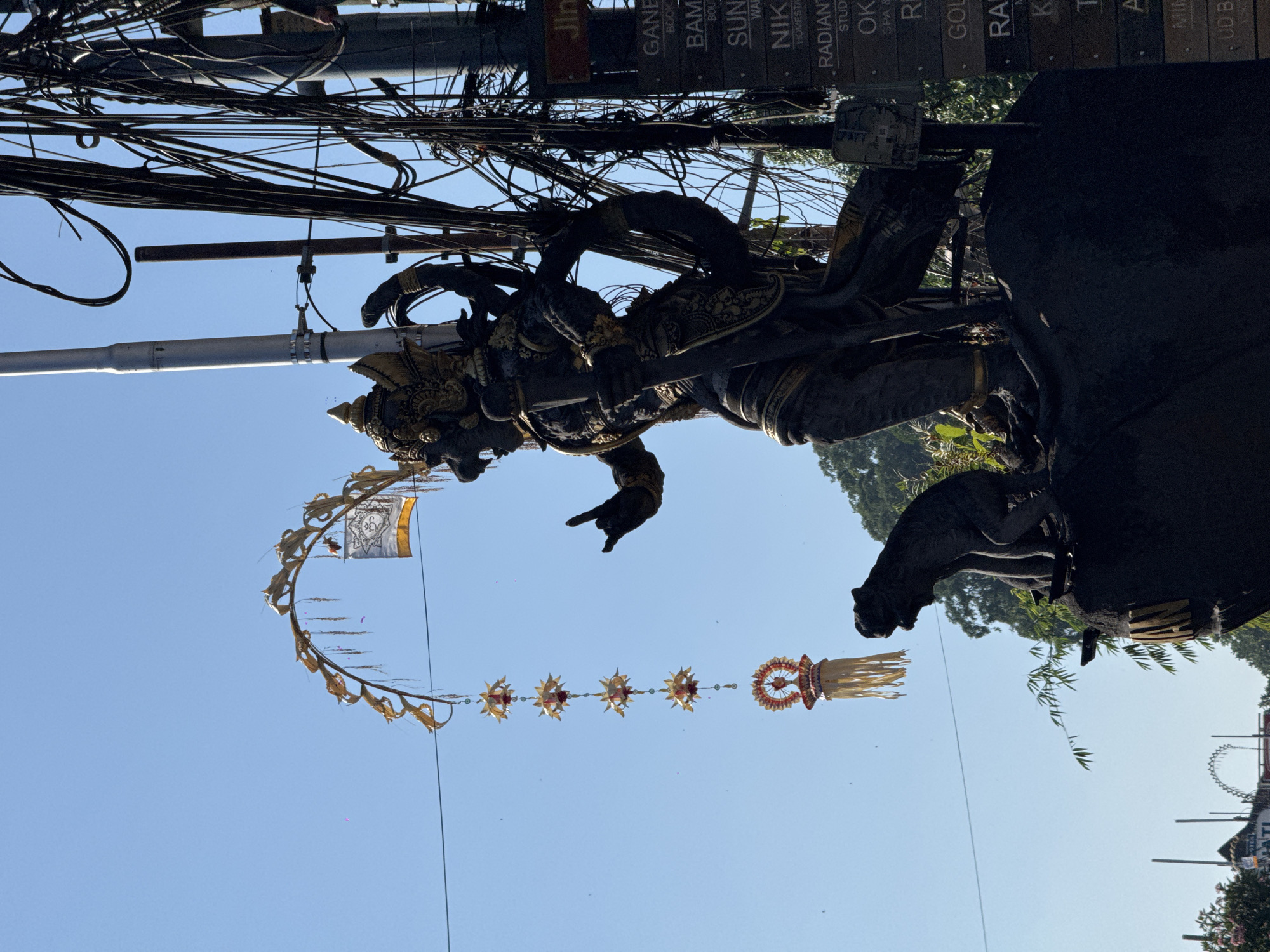 A dark statue of a multi-armed deity with ornate golden headwear stands in front of a bright blue sky in Ubud, Indonesia. The statue is adorned with golden trim and holds a weapon in its right hand. To the left of the deity, a smaller dark statue of a monkey sits on a rock. Hanging from a pole in the sky are several golden star-shaped ornaments and a decorative banner. The background features a dense network of electrical wires and a street sign with multiple listings, including "GANES," "BAM," "SUN," "NIK," "RADIANT," "OK," "RA," "K," "T," and "A." The lower portion of the image shows lush green foliage and trees.