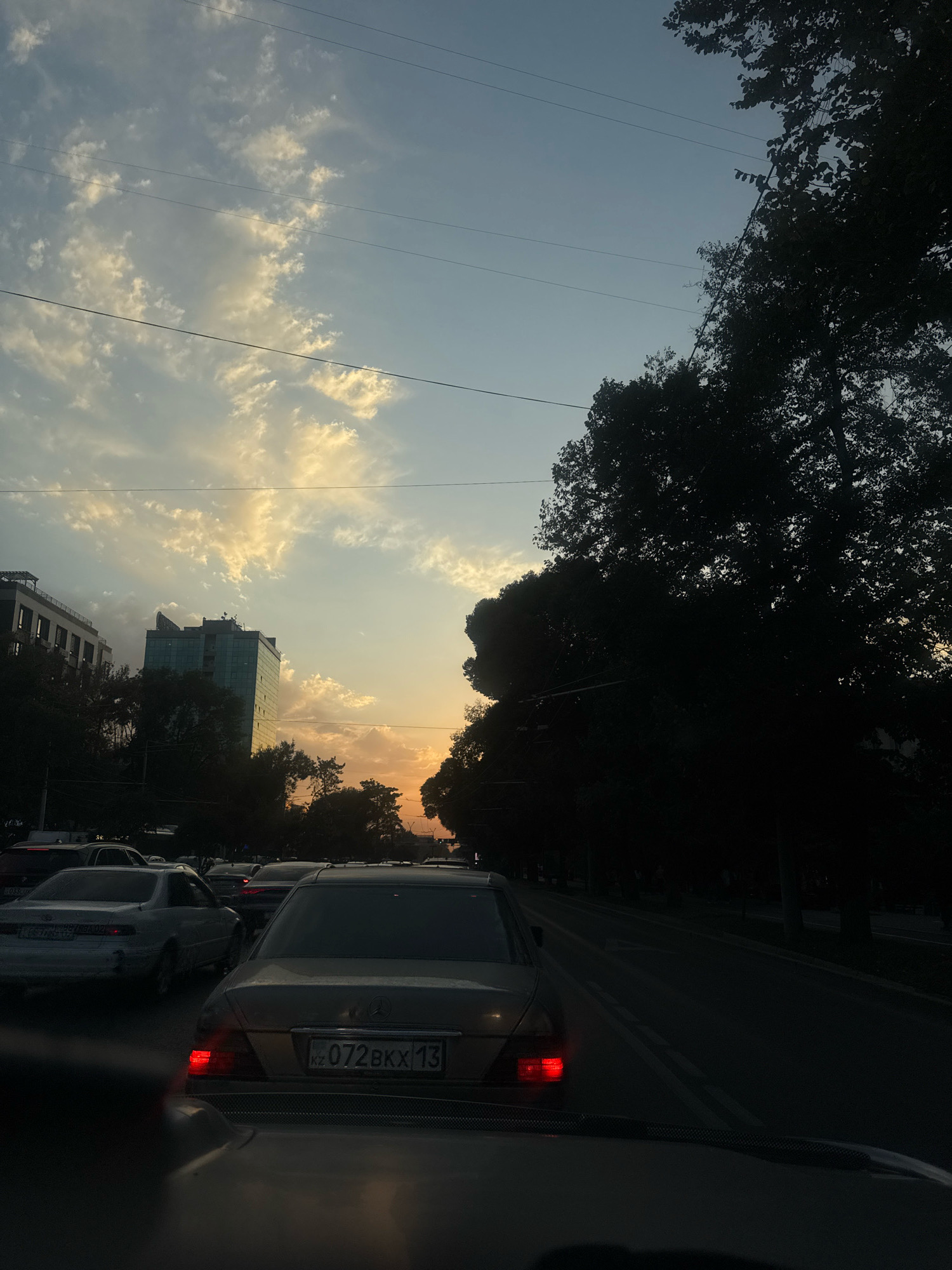 The image captures a street scene in Almaty, Kazakhstan, during a sunset. Traffic is heavy, with several cars visible on the road. In the foreground, the back of a light-colored sedan is prominent, featuring a visible license plate with the inscription "072BKX13". To the left, another white sedan is seen in traffic. In the background, a modern glass-fronted building stands out among the trees, and the sky is illuminated with soft hues of orange and yellow from the setting sun, with scattered clouds. The overall atmosphere suggests a typical evening rush hour in a city.