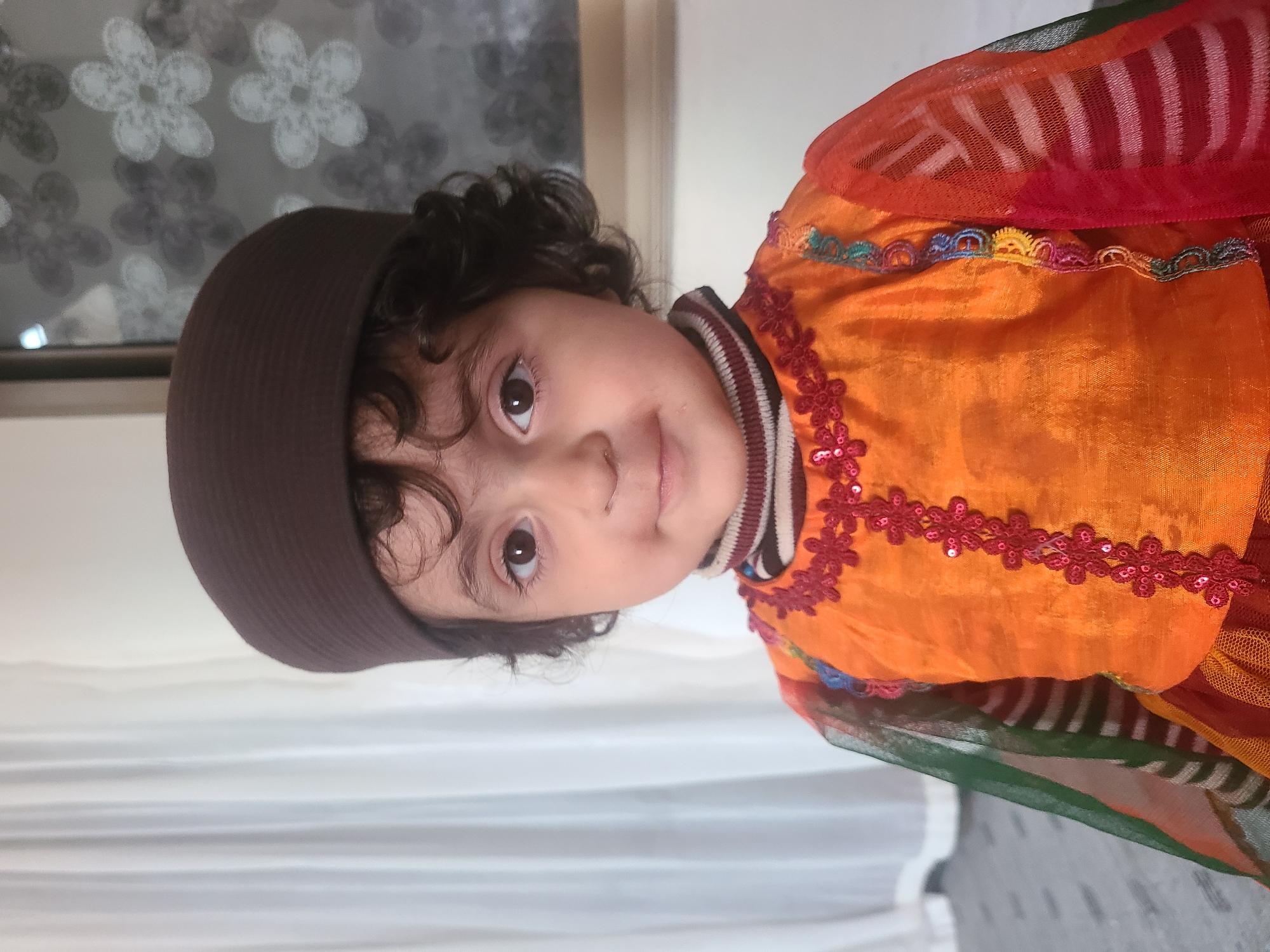 A child wearing a brown hat and an orange and red outfit with floral embellishments is visible. The background includes a patterned window and white drapes. The location is likely Quetta City Tehsil, Pakistan.