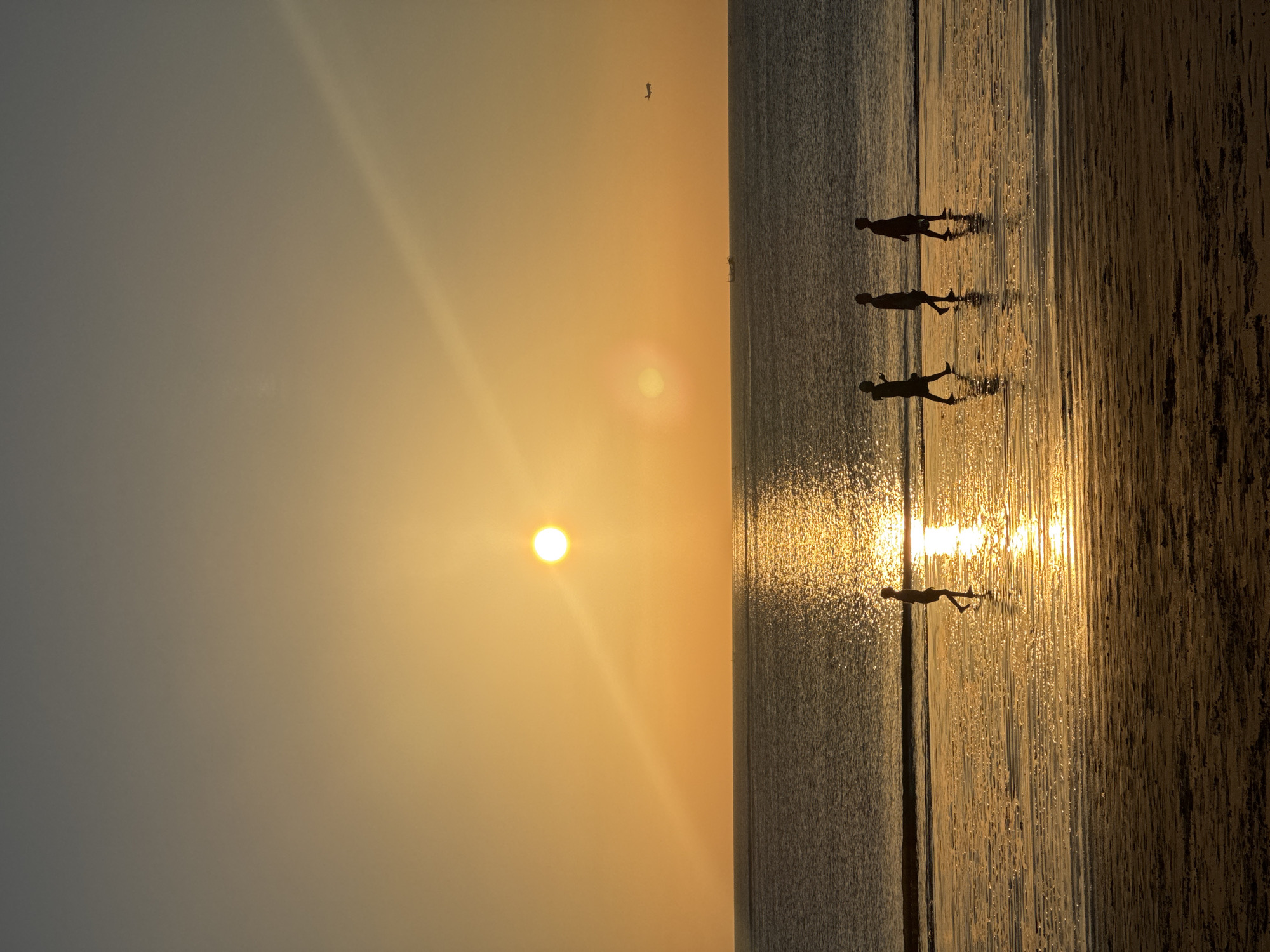 A vertical, full-frame photograph captures a serene sunset over the ocean at Kuta Selatan, Indonesia. The sun, a bright, circular orb, hangs low in the sky, casting a golden glow across the water and the wet sand of the beach. A narrow, diagonal sunbeam emanates from the sun, stretching across the frame. Four silhouetted figures, appearing as children, are walking along the shoreline, their forms stark against the shimmering water. The ocean is calm, with gentle waves lapping at the shore. In the distance, a faint airplane can be seen in the sky, and a small boat is visible on the horizon. The sand in the foreground exhibits a textured, rippled pattern, reflecting the warm light of the setting sun.
