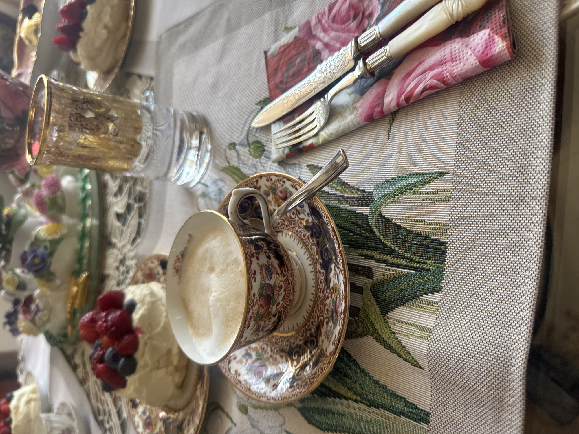 The image depicts a close-up of a table setting, likely for afternoon tea or a light dessert. The foreground features a cup of coffee with frothy milk, placed on a saucer with an intricate floral pattern. To the right of the cup, a set of silverware, a fork and a knife, rests on a floral-patterned napkin. The silverware has ornate handles, suggesting it is vintage or fine quality. A small, individual dessert, possibly a meringue or cake topped with fresh berries like strawberries and blueberries, sits on a decorative plate to the left of the coffee cup. In the background, a glass with a golden, embossed design and another small dessert are visible. The table is covered with a placemat that has a textured, neutral base and a prominent green and white botanical pattern. The overall impression is one of an elegant, perhaps slightly formal, occasion. The scene is set on a table, possibly indoors or in a shaded outdoor area, with the lighting suggesting daytime. There are no people visible in the image. The image does not contain any information that would allow for the identification of its specific location within the city of Plovdiv, Bulgaria.