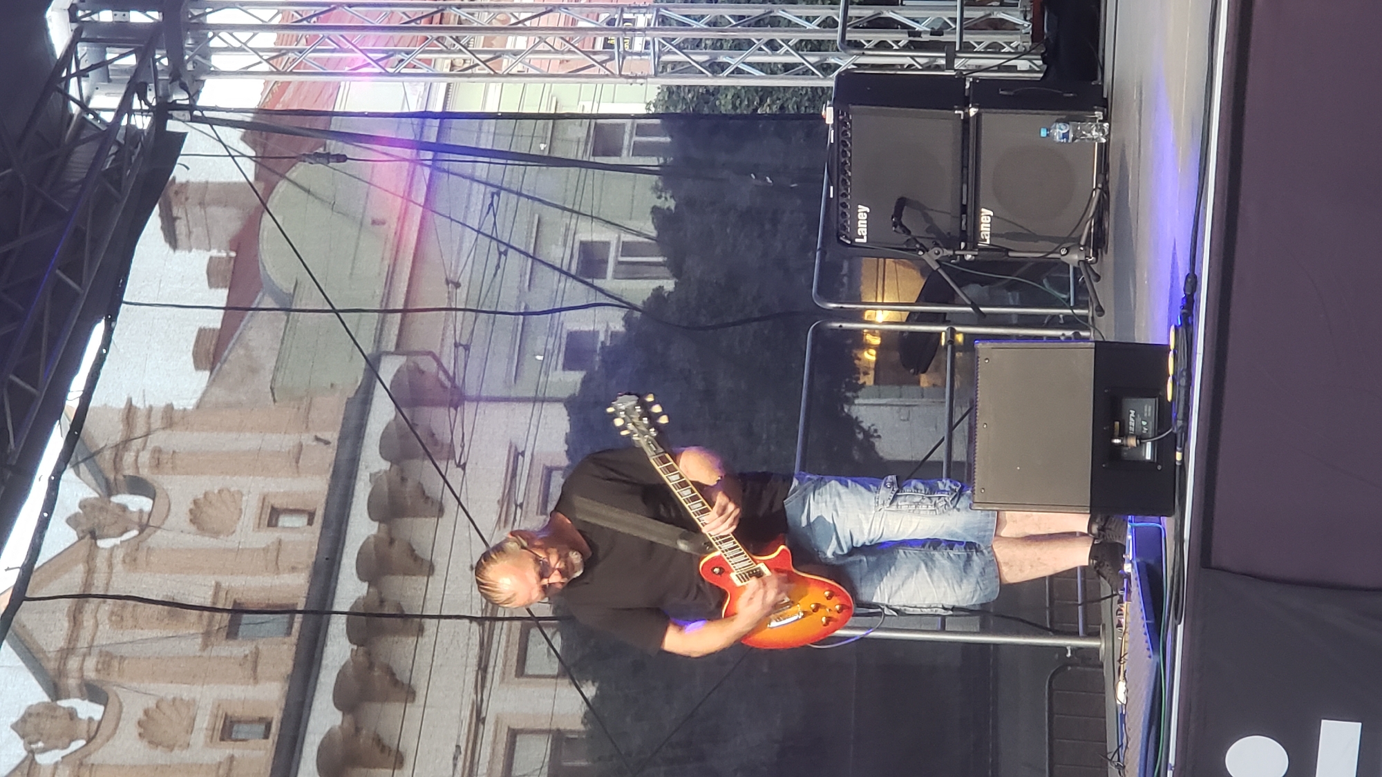 A man is playing an electric guitar on a stage in Prešov, Slovakia. He is wearing a black t-shirt, denim cargo shorts, and black sneakers. The guitar is red and orange. Behind him, there is a black backdrop with an image of buildings. To his right are two Laney speaker cabinets. The stage is set up outdoors, with a metal truss structure visible behind the backdrop. The buildings in the backdrop appear to be in an older architectural style, possibly indicative of a historic city center. The lighting on stage is a mix of natural daylight and stage lighting, with some purple and blue hues visible.