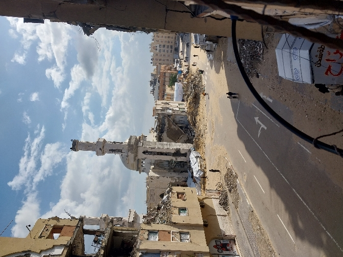 This image depicts a street scene in Khan Yunis, Palestinian Territory, under a partly cloudy sky with strong sunlight casting shadows. The perspective is from an elevated position, looking down a main thoroughfare flanked by extensively damaged buildings.
On the left, a multi-story building stands with significant structural damage, its facade partially collapsed, revealing exposed interiors and rebar. Large piles of rubble accumulate at its base. In the midground, a prominent mosque with a minaret and dome is severely damaged; the minaret remains partially intact but shows visible signs of distress and debris at its base, while the main mosque structure is largely reduced to rubble. Beyond the mosque, more damaged buildings and debris piles are visible, extending towards a background of less severely affected, multi-story urban structures. On the immediate right, the frame is partially obscured by another damaged building, with hanging wires or cables in the foreground.
The paved road running through the center features white lane markings, including a dashed line, a solid line, and a white left-turn arrow. The road surface is dusty with small debris. Large accumulations of building rubble and dirt line both sides of the street.
Several individuals are present on the street. Two people walk together in the midground on the left side of the road, casting long shadows. Another individual stands near a bicycle parked against a pile of rubble on the left. Multiple other figures are visible walking further down the street in the distance. In the lower right corner, a partially visible white structure, possibly a temporary kiosk or sign, displays Arabic text, including "بطة شحن" (battery charging) and "زواج" (marriage). A flock of birds flies in the upper right portion of the sky.