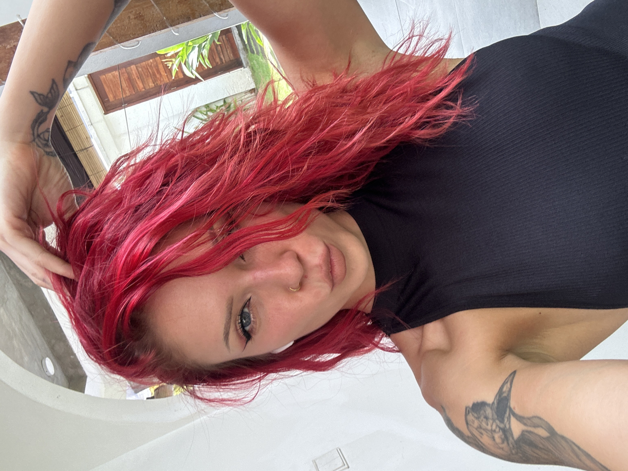 A woman with red hair and tattoos is posing for a selfie inside a building, likely located in North Kuta, Indonesia. She is wearing a black top. The background includes a glimpse of the outdoors through a window with wooden blinds and some foliage.