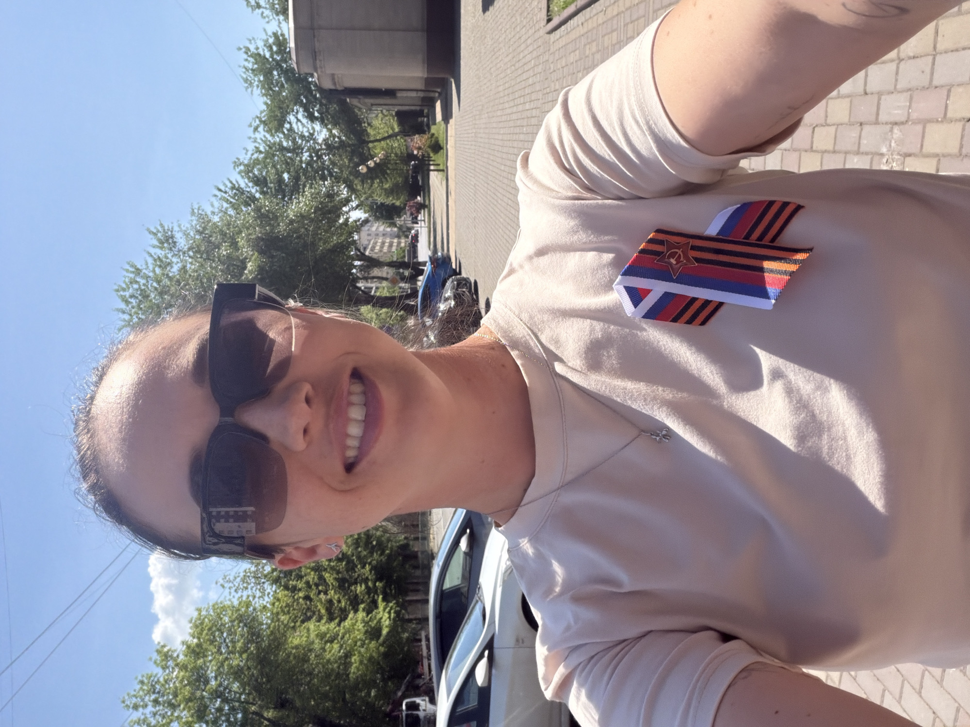 A woman is taking a selfie outdoors. She is wearing sunglasses and a t-shirt with a ribbon pinned to it, possibly a St. George ribbon repurposed with the Russian tricolor and a red star emblem. The background shows a city scene with trees, buildings, and parked cars. Based on the emblem on the ribbon, it could be inferred that this image was taken in Russia. Since the model does not provide explicit information about the location as Grozny, Russia, it can not be confirmed, however there are signs of Eastern European or Russian architecture visible.