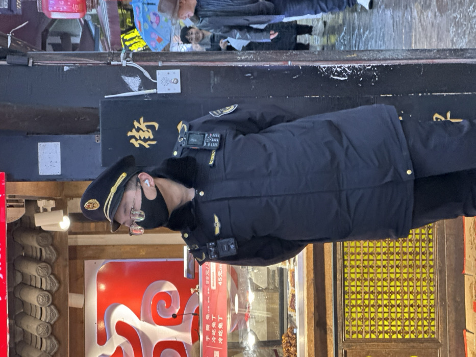 A uniformed security officer in a black hat and face mask stands in front of a storefront with red signage advertising "手撕兔" (hand-shredded rabbit) for 88 yuan per unit, and "冷吃兔丁" (cold spicy rabbit cubes) priced at 45 yuan. Above, weathered black paint and faded calligraphy details the building exterior. In the background, blurred figures and reflections can be seen in a window.