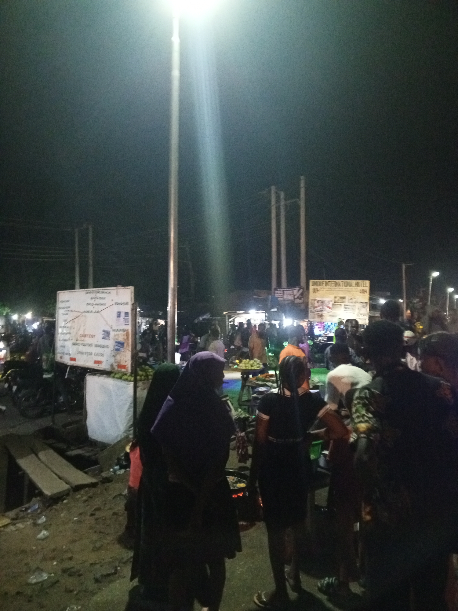 At night in Unknown, Unknown, a bustling street scene unfolds. Lit by bright overhead streetlights, a crowd of people, including a woman in a purple hijab, clusters around various stalls and vendors. Signboards and advertisements are visible in the background, along with parked motorcycles, adding to the urban ambiance. The ground is uneven and littered with debris.