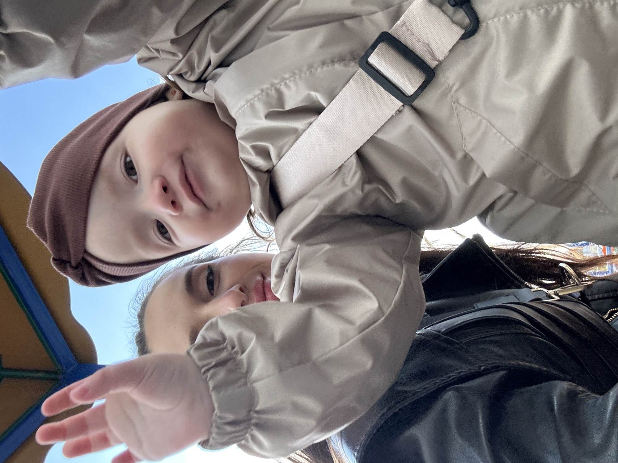 This media file captures a playful, low-angle moment outdoors, with the image rotated approximately 90 degrees counter-clockwise from a standard upright orientation. The scene is set against a bright, clear blue sky, suggesting a sunny day in an outdoor location, potentially a park or playground in Yekaterinburg, Russia.
The primary subjects are a young child and an adult, both looking down towards the camera with gentle expressions. The child, positioned higher in the frame (appearing towards the top left of the current orientation), is a toddler or preschooler wearing a brownish knit hat or headband and a light beige, padded jacket. A light-colored strap, possibly from a carrier or bag, runs diagonally across the child's chest. The child looks directly at the camera with a subtle smile.
Below and partially obscured by the child, an adult's face (eyes and nose) is visible, also looking towards the camera. The adult is wearing a dark jacket, possibly leather, and has their right hand extended downwards and slightly towards the camera. Part of a blue and yellow structure, possibly playground equipment, can be seen on the far left edge of the image.
The overall mood is warm and engaging, highlighting a tender interaction between the individuals. There is no visible text within the image.