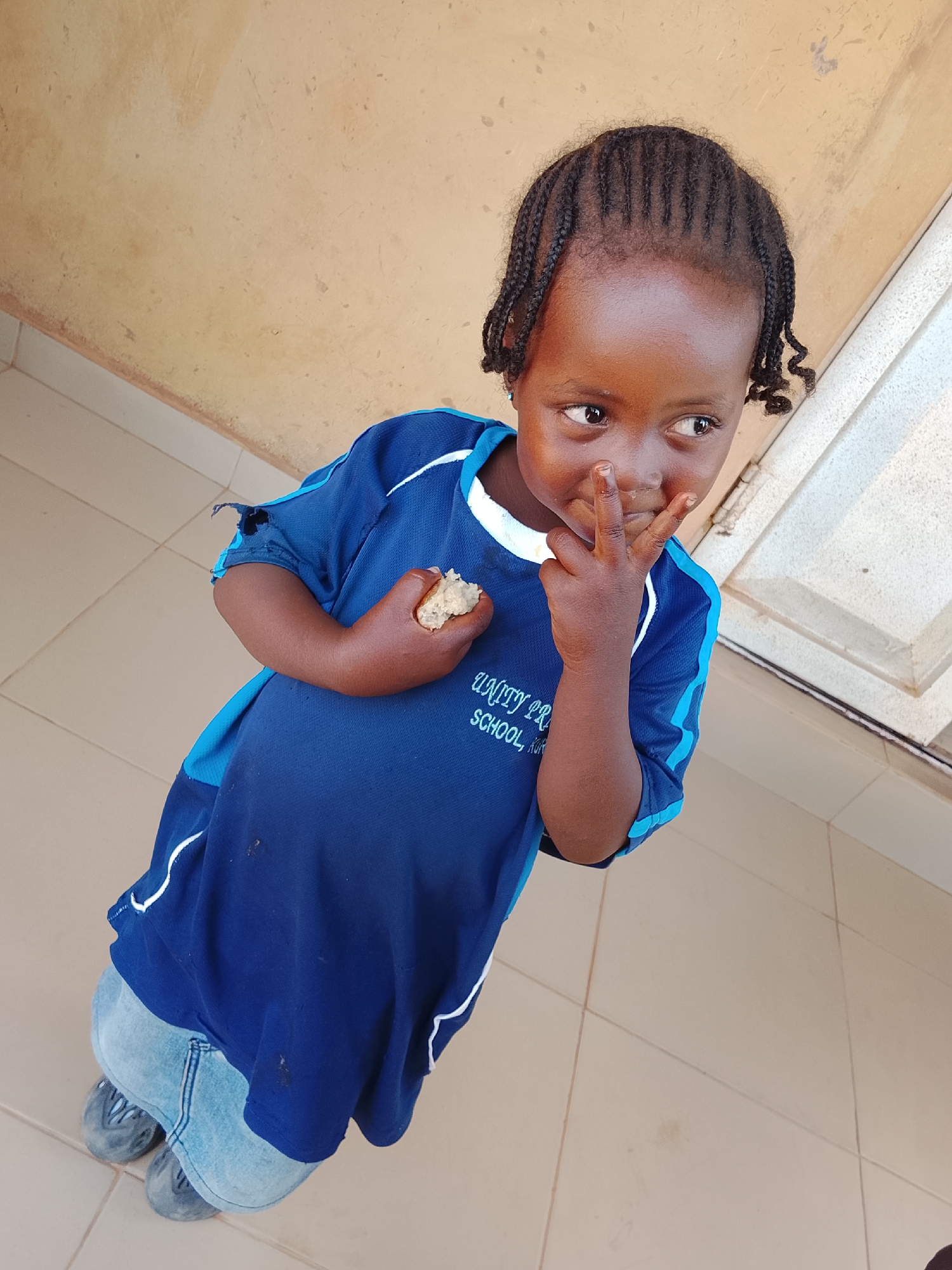 A young child stands on a tiled floor, likely indoors, wearing a blue shirt with "UNITY PR SCHOOL, KUR" printed on it, and jeans. The child holds a piece of bread in one hand and has two fingers raised to their face in the other hand. The setting suggests a location in Kuru Dungus, Nigeria, implied by the text on the shirt. The child is looking off to the side with a slight smile.