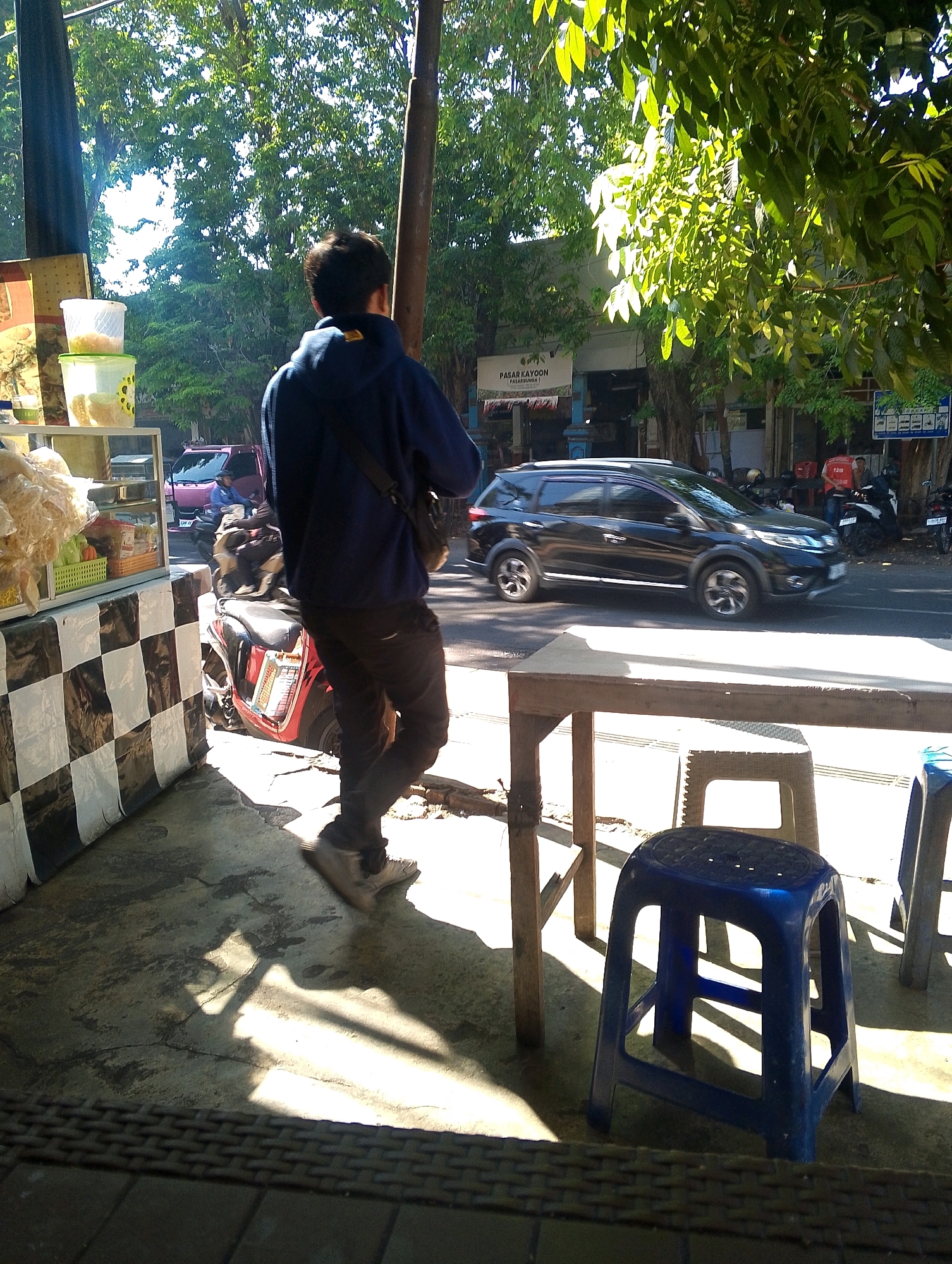 The image shows a street scene with a person standing in the foreground, facing away from the camera. They are wearing a dark blue hooded jacket and black pants. To the left, there is a stall with shelves containing food items and white plastic containers. A red motorcycle is parked next to the stall. In the background, a black SUV drives past on a road lined with trees and buildings. A sign that says "PASAR KAYOON" is visible on one of the buildings. Several motorcycles are parked on the side of the road, and a few people are walking. The scene appears to be a street market or a roadside stall in a city. The lighting suggests it is daytime, with strong sunlight creating shadows. There are also two blue plastic stools and a wooden table in the foreground.
