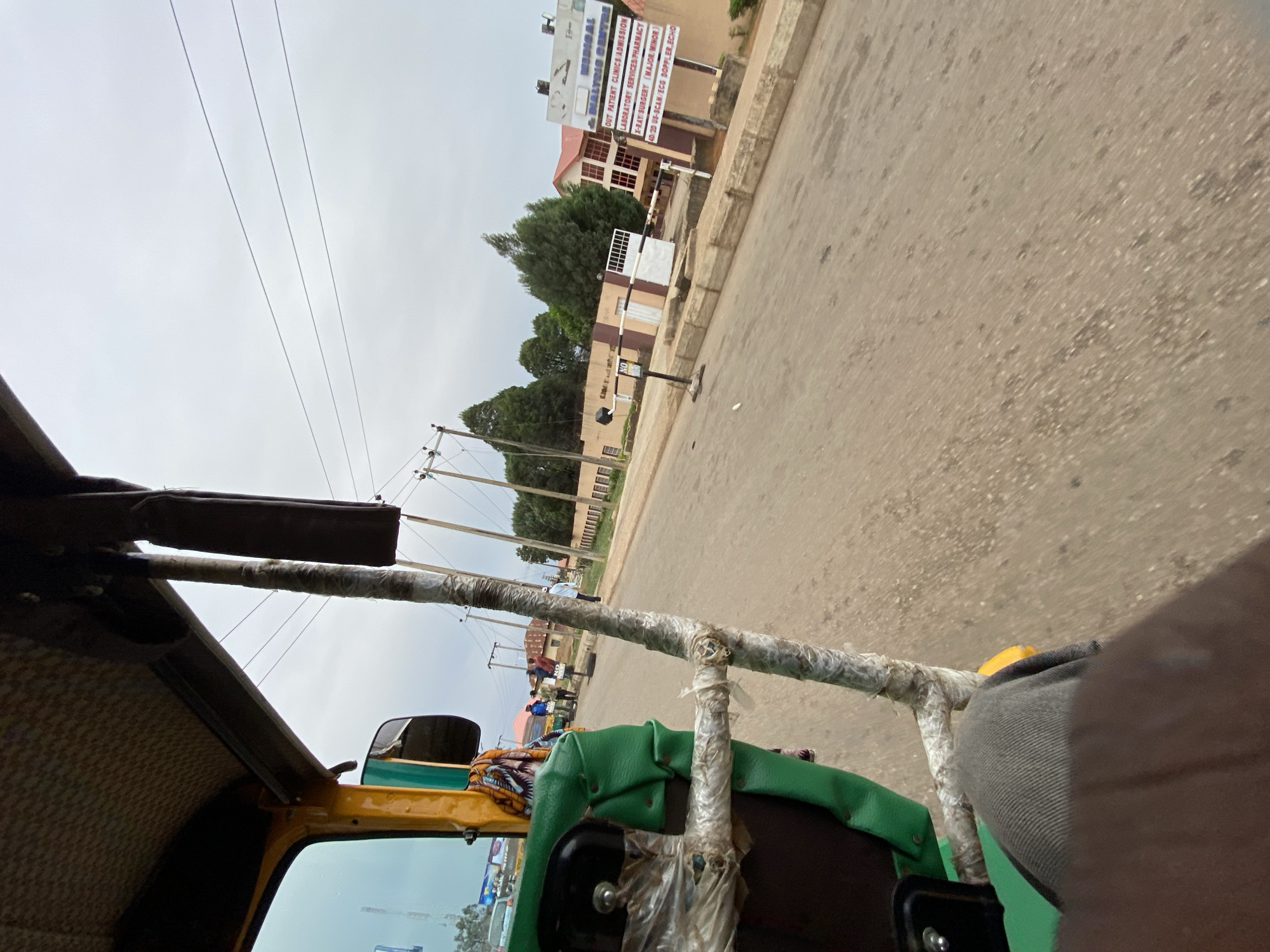 The image is a first-person perspective shot from inside a vehicle, possibly a tricycle or auto-rickshaw, moving along a paved road in what appears to be a residential or commercial area. The view is from the passenger seat. The road is mostly empty, with a few distant pedestrians and parked vehicles visible on the left side of the road. Utility poles with power lines run along the left side of the road.
A building with signage indicating "NALYSI MEDICAL CENTER" is visible in the background. The sign lists services like "OUT PATIENT CLINICS ADMISSION, LABORATORY SERVICES PHARMACY, X-RAY SURGERY (MAJOR/MINOR), 4D/2D US-SCAN/ECG DOPPLER, ECHO." The sky is overcast.
Based on the medical center and available information, it is highly probable that the location is in Jos, Nigeria.