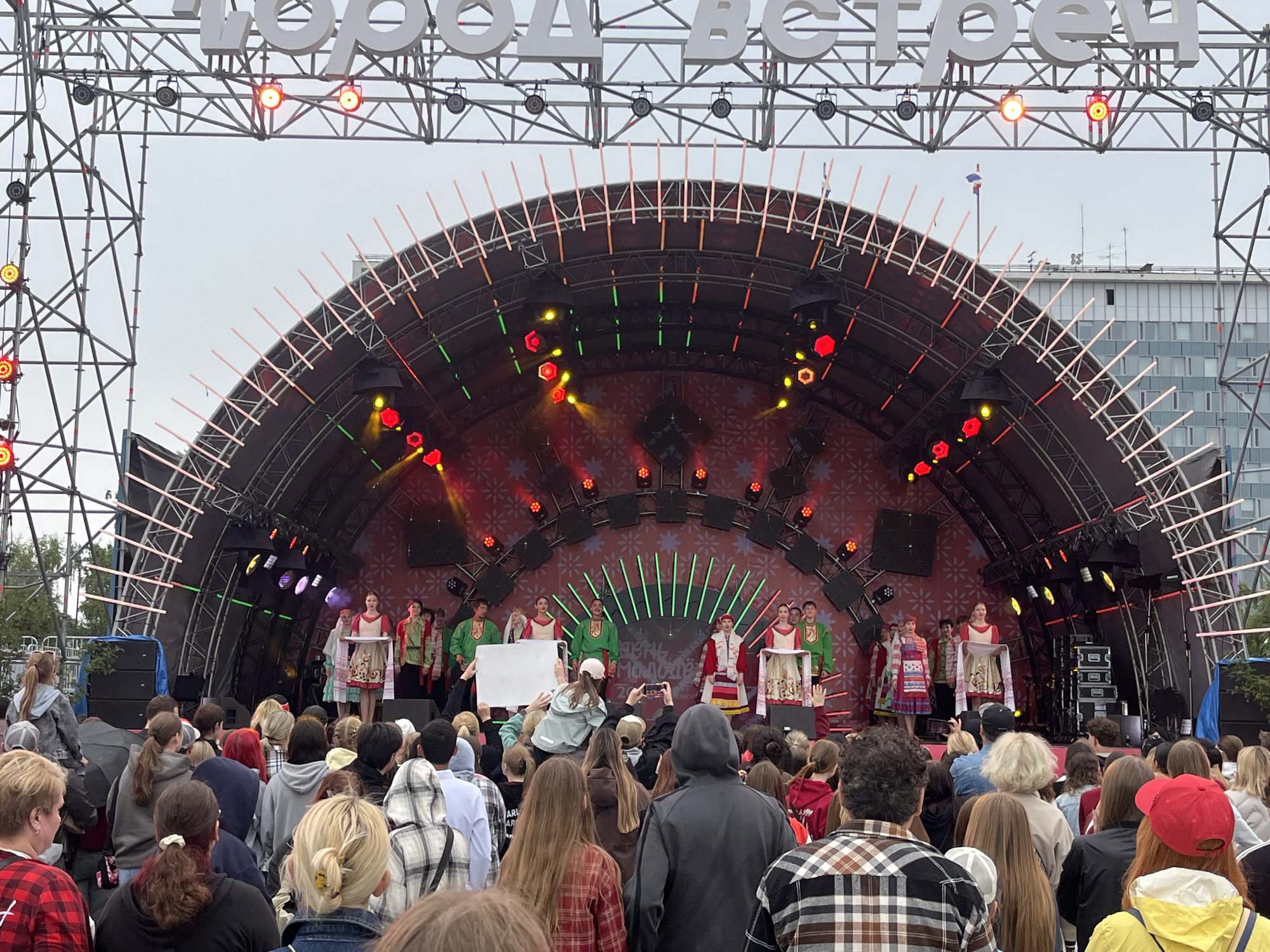A large outdoor stage is set up for a performance. The stage features a curved backdrop with a red pattern and green vertical lights. Several people, dressed in traditional colorful clothing, are on stage. Some are holding a white banner, while others are standing by a table with a white cloth. A young woman in a red and white dress stands center stage, holding a tray or platter. A crowd of spectators is gathered in front of the stage, many of them young people wearing casual clothing. The weather appears overcast, suggesting it might be morning or late afternoon. The overall scene indicates a cultural event or festival taking place in Perm, Russia, likely a performance celebrating local traditions. The large white letters at the top of the stage structure read "ГОРОД ВСТРЕЧ" which translates to "City of Meetings."