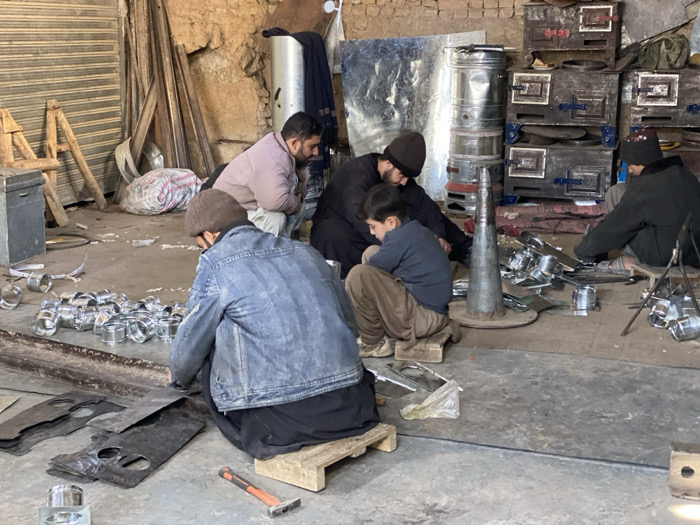 The image depicts an interior view of a metalworking workshop in Kandahar, Afghanistan. Five male individuals, ranging from children to adults, are engaged in manual fabrication tasks on a concrete floor.
The workshop environment features rough, unplastered walls, with a corrugated metal roll-down shutter visible on the left. In the background, raw materials including metal rods and a bundled sack of scrap metal are present. Several large, dark-colored metal stoves with rectangular doors are stacked, alongside a tall, shiny cylindrical metal tank and a prominent conical metal form.
In the immediate foreground-left, a male individual wearing a brown beanie and a blue denim jacket squats on a wooden stool, manipulating flat, dark metal sheets, some exhibiting pre-cut circular holes. A hammer with an orange-black handle lies on the floor next to him. To his right, an adult male in a light-colored jacket squats, observing an accumulation of small, shiny cylindrical metal components.
Centrally, an adult male in dark clothing and a dark beanie squats, appearing to supervise a male child positioned to his immediate left. The child, wearing a dark blue top and light brown trousers, also squats on a wooden stool, focused on metal pieces. Further right, another adult male, dressed in dark attire and a dark beanie, squats while actively working with metal components in proximity to the stacked stoves. The scene indicates ongoing manual assembly and fabrication of metal products, utilizing numerous small metal parts distributed throughout the workspace.