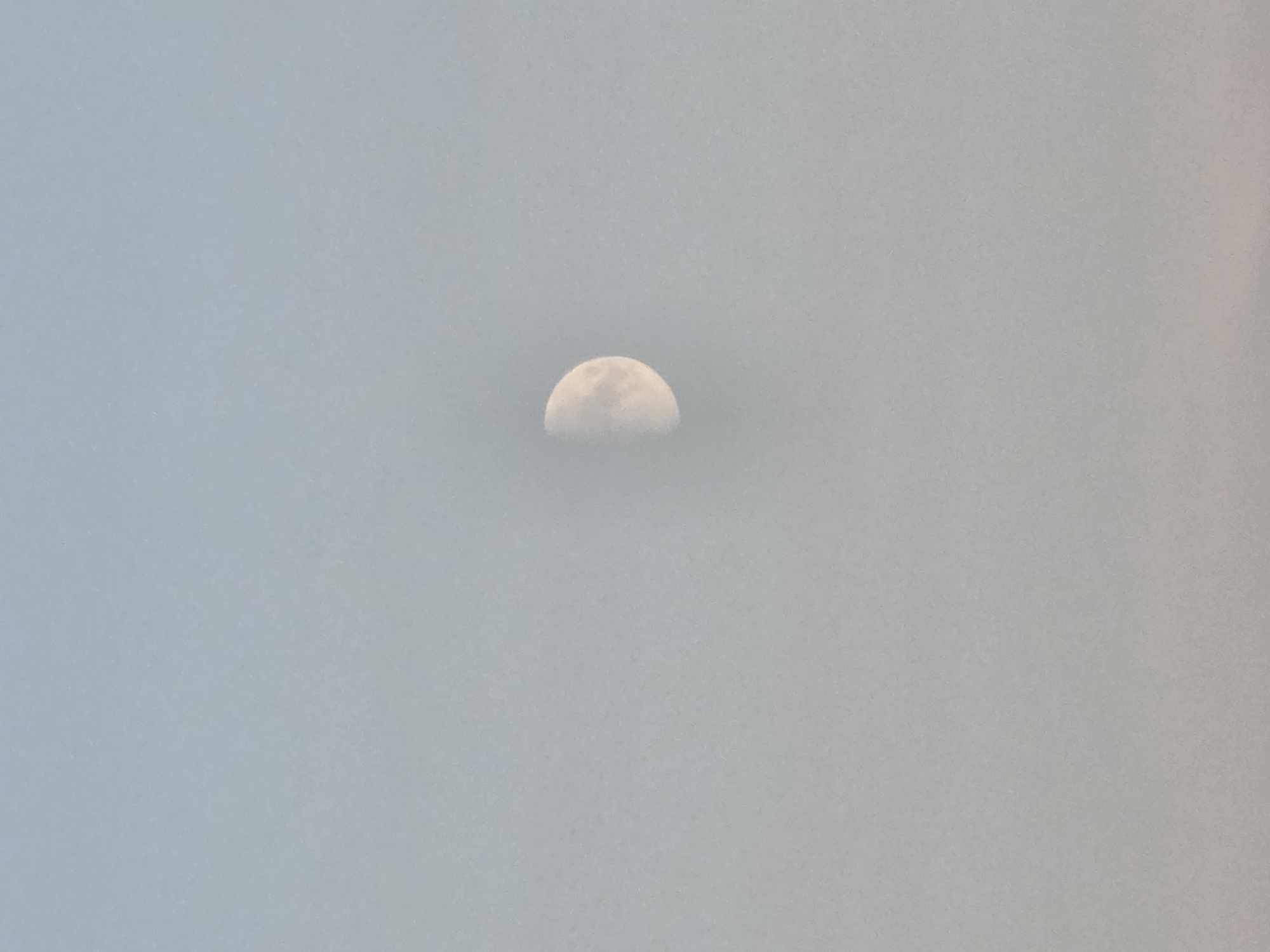 The image is a photograph of the moon in a hazy sky. The moon is approximately half-illuminated and appears slightly blurred due to atmospheric conditions. The sky is a pale blue-gray, with a subtle gradient of lighter tones towards the bottom of the frame. There is no discernible indication of the city of Речицы, Russia, or any specific urban features present in the image. The scene is devoid of any people, objects, or actions.