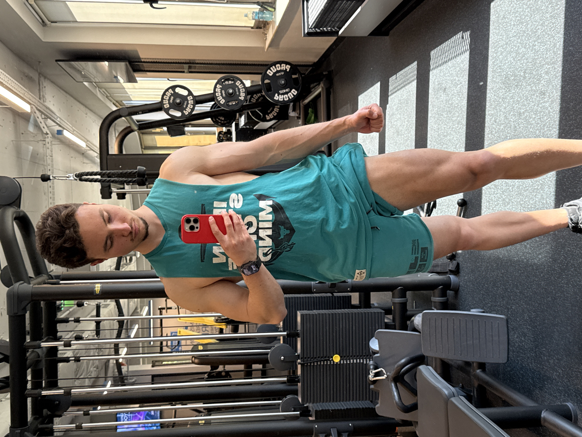 A fair-skinned man in athletic attire poses for a photograph within a fitness facility. He is wearing a green tank top and shorts, holding a red smartphone, and sporting a black smartwatch. He stands beside a weightlifting machine, which is set against the backdrop of gym equipment and window light. The location is consistent with the interior of a modern gymnasium, but the precise city and country (Sofia, Bulgaria) are not discernible from the visual data provided.