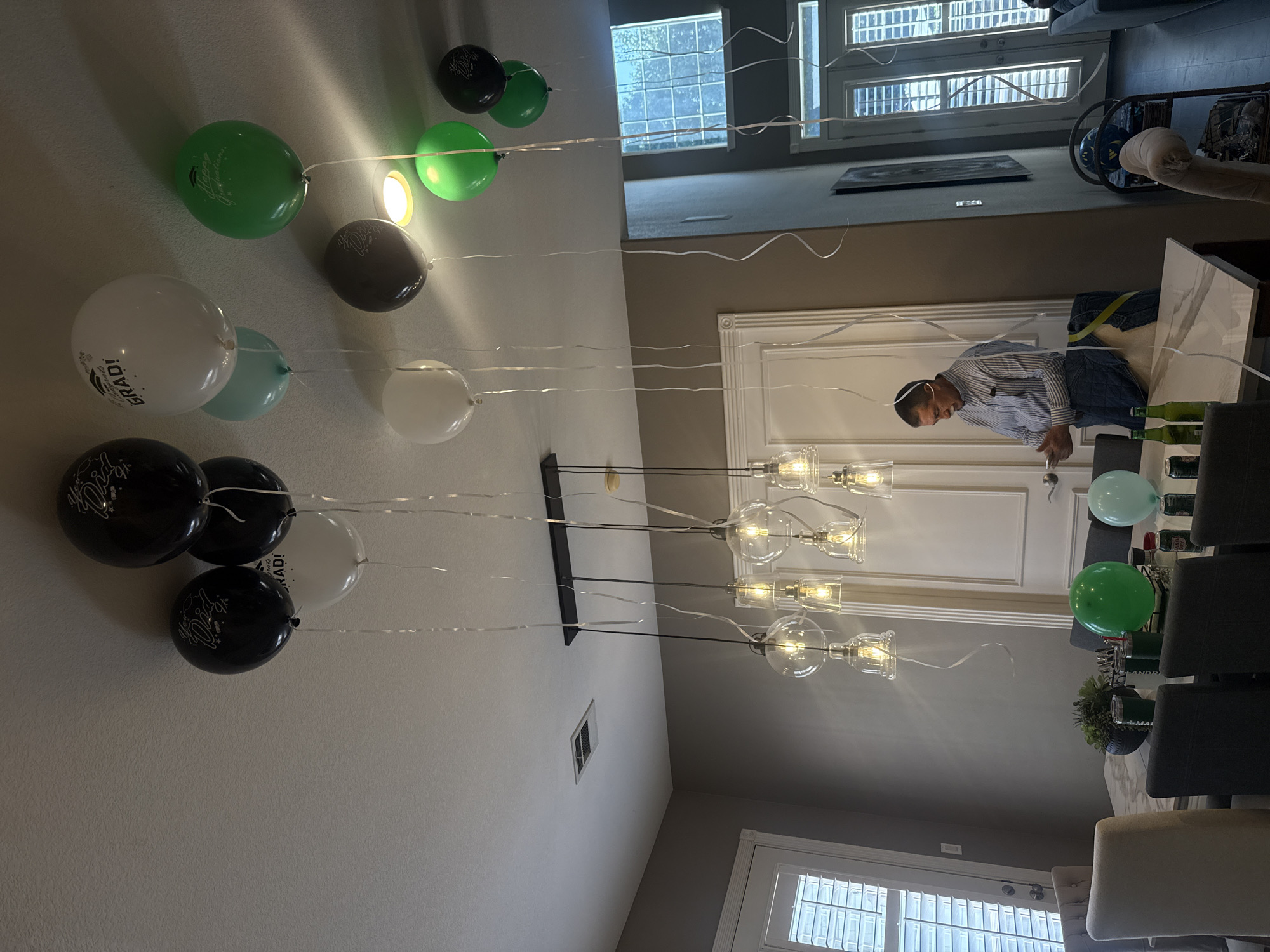 A residential interior in Southlake, Texas, United States of America, shows a person at a dining table with chairs, bottles, and balloons. The scene includes a multi-bulb light fixture, windows, and a decorative wall trim. Various balloons float overhead, some with graduation motifs. The person is dressed in a striped shirt and jeans and appears to be interacting with objects on the table. The overall environment suggests a celebratory gathering.