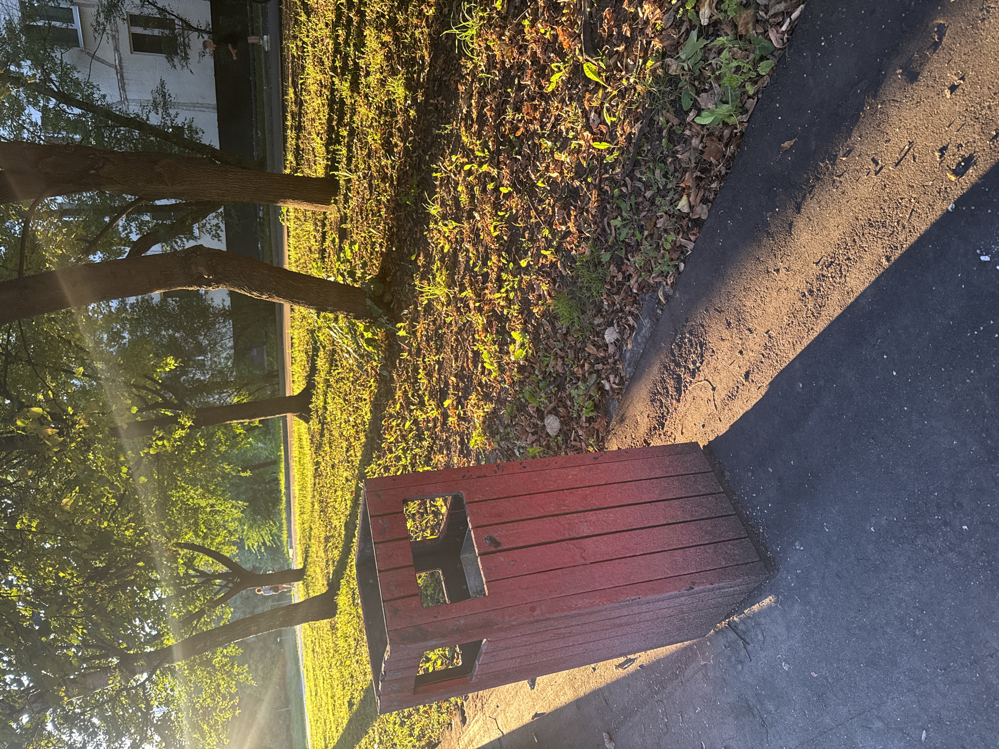 A red, rectangular trash receptacle stands on the side of an asphalt path. The trash can is made of vertical wooden slats and has two square openings on its front. The ground around the trash can is covered with dry leaves and sparse green vegetation. Several trees with full green canopies are visible in the background, with sunlight filtering through their leaves and casting long shadows on the ground. In the distance, a building with white walls and dark windows can be seen. A person is walking in the background on the left side of the image. The scene appears to be a park or residential area in Moscow, Russia, likely during the late afternoon due to the long shadows and golden light.