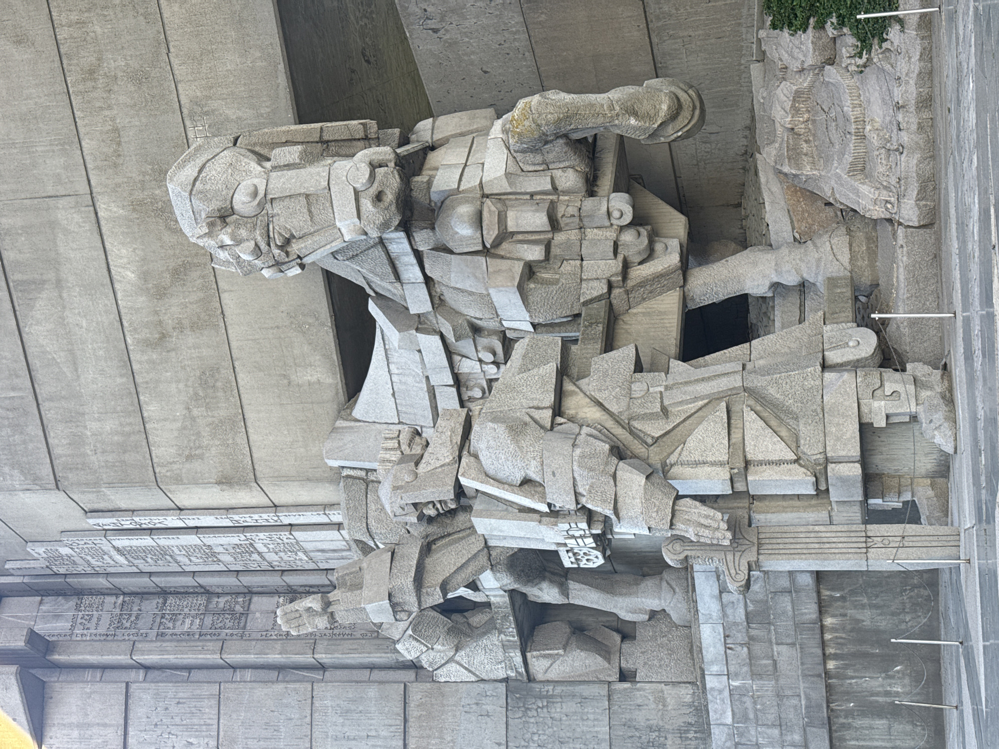 The image captures a detailed view of a monumental concrete sculpture located at the Monument to 1300 Years of Bulgaria in Shumen, Bulgaria. The sculpture is highly stylized and depicts a heroic figure, potentially a soldier or warrior, rendered in an abstract, cubist manner using large, geometric blocks of concrete. The figure is portrayed in a dynamic pose, seemingly in action or defense. The sculpture is mounted on a large concrete structure. There are also sections of the building with inscribed texts. The scene is bathed in daylight, emphasizing the concrete texture. The sculpture is part of a larger complex commemorating the 1300th anniversary of the First Bulgarian Empire.