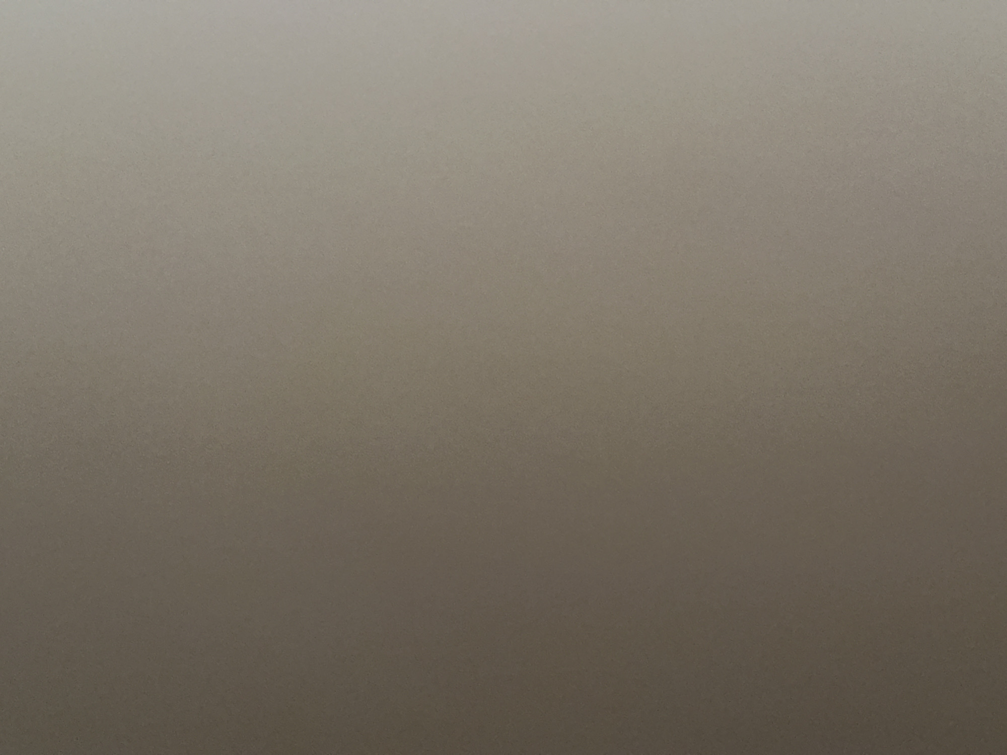 This is a full-frame, medium shot photograph of a plain, textured surface. The image is predominantly composed of a gradient of grey tones, transitioning from a lighter shade at the top to a darker shade at the bottom. The texture of the surface appears to be slightly grainy or matte, with subtle variations in light and shadow that suggest a non-uniform material. There are no discernible objects, people, or any specific scene or setting present in the photograph. The lighting appears to be even and diffused, with no strong shadows or highlights, making it difficult to determine the time of day or specific environmental conditions. Similarly, there are no location cues visible, and no text is present in the image. The overall impression is one of minimalism and abstraction, focusing solely on the texture and color of the surface.