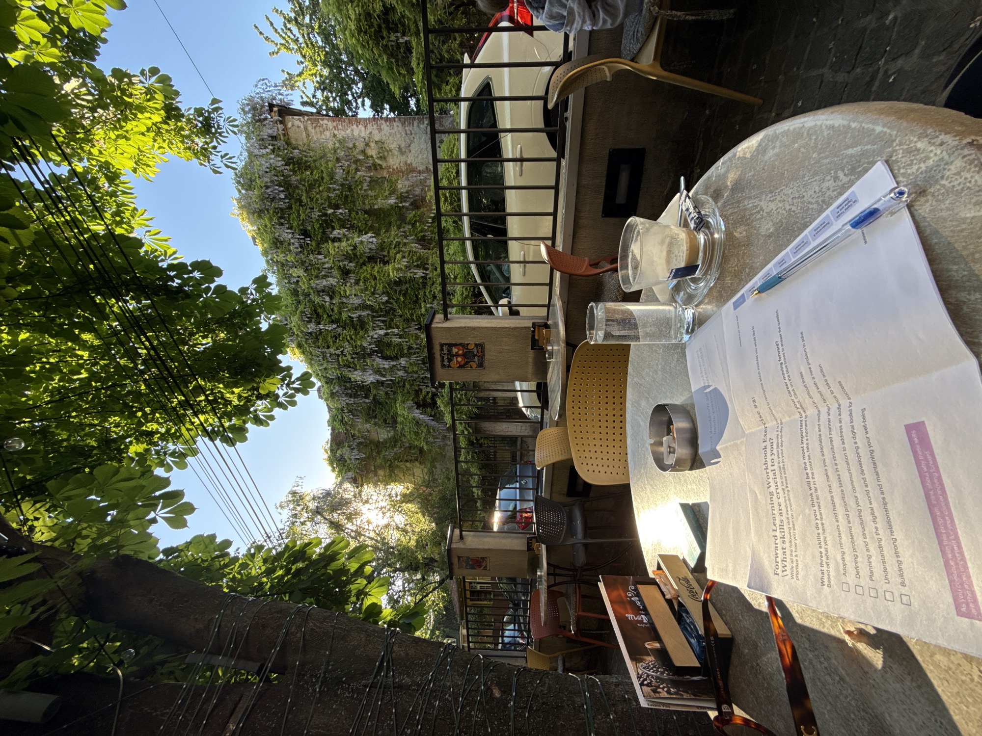 The image depicts a cafe scene in the Vracar Urban Municipality, Serbia. The shot is taken from a table, looking out onto the street.
On the table, there is a used coffee cup, a glass of water, an ashtray, books, a pair of glasses, a pen, and a worksheet titled "Forward Learning Workbook Exercise #2." The worksheet asks the reader to reflect on skills that will be crucial to their journey.
Through a wrought iron fence, a white car is parked on the street. A building covered in wisteria vines is visible behind the car.
The scene is bathed in sunlight, and the branches of trees provide shade over the cafe. A person is partially visible at the top of the image. Other tables and chairs are visible in the background.