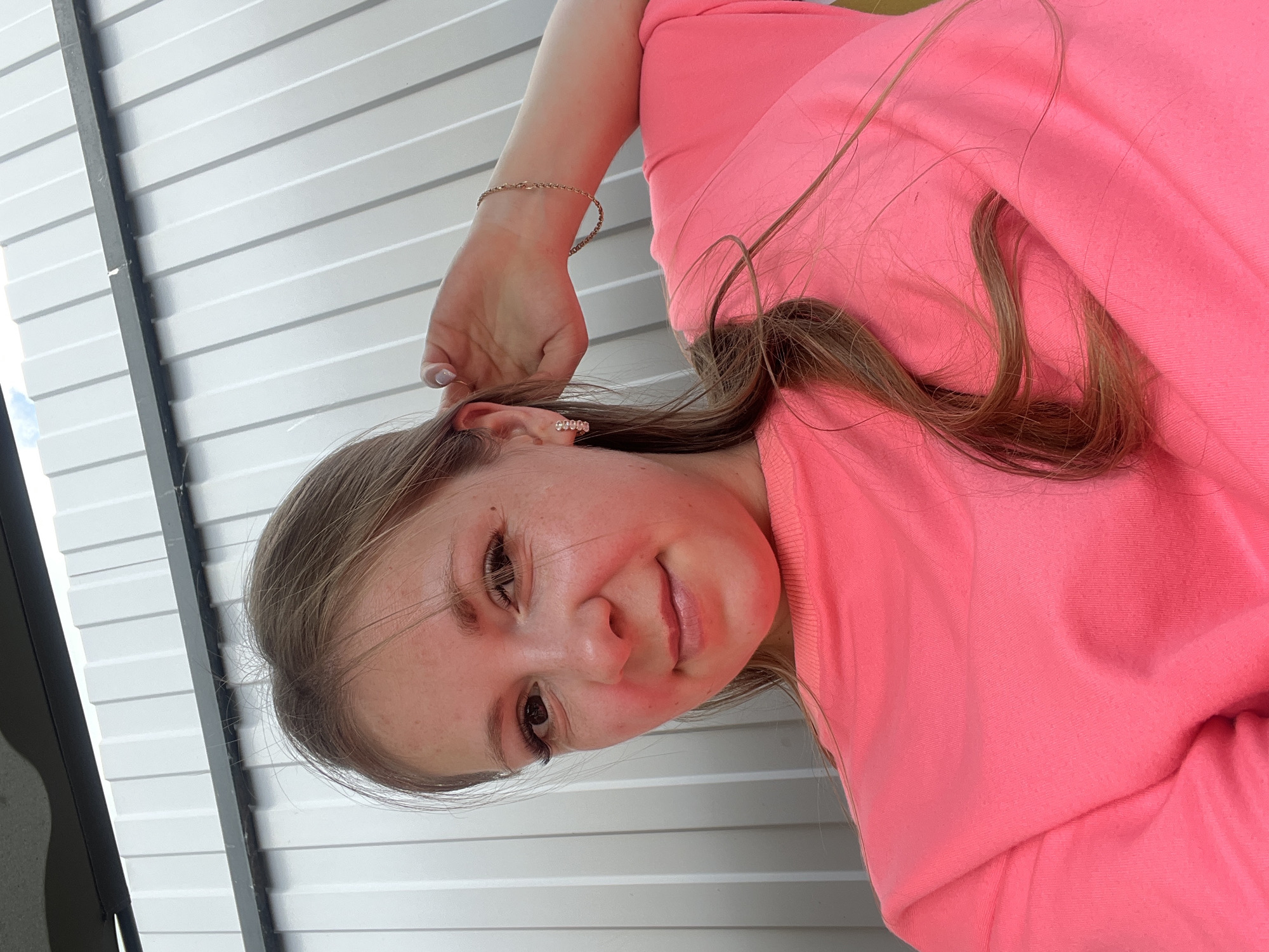 A young woman is taking a selfie outdoors. She is wearing a bright pink sweatshirt and a bracelet on her right arm. Her hair is light brown and she has earrings in both ears. The background is a corrugated metal wall. The image appears to have been taken during the day, and the lighting suggests an outdoor setting, possibly in the city of Sukhyrinskoye, Russia, though no specific landmarks are visible.