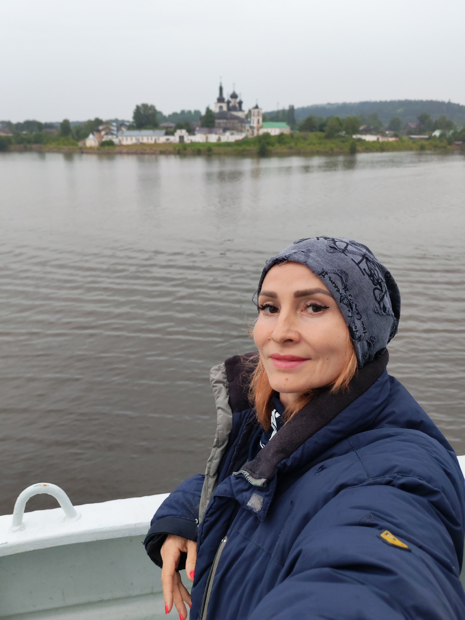 In the foreground, a woman with auburn hair and makeup is captured in a selfie from the chest up. She is wearing a dark blue winter jacket and a grey beanie with a pattern. The woman is standing on the deck of a vessel, as indicated by the white railing and a white loop fixture in the lower left corner of the image. In the background, across a body of water, the Goritsy Monastery is visible, a prominent landmark in Goritsy, Russia. The monastery features several white buildings with dark domes and a prominent bell tower. The environment appears overcast and the water is calm.
